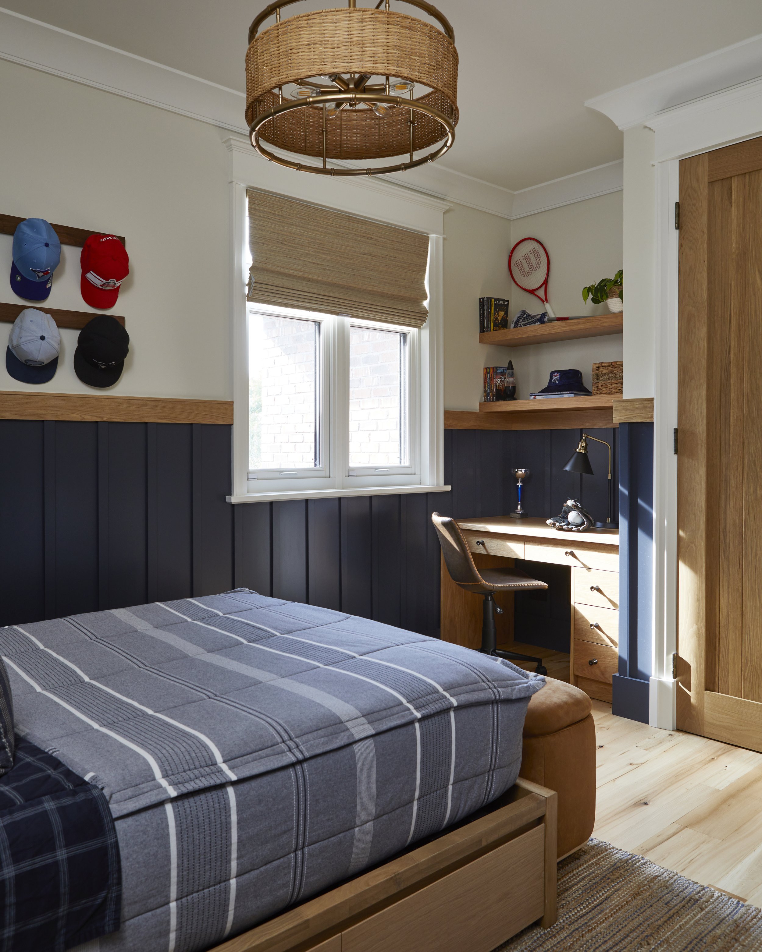 A cozy bedroom featuring a bed with a blue plaid mattress, a desk near a window with a brown chair, books, and hats on wooden shelves, window blinds, and a round wicker ceiling light.