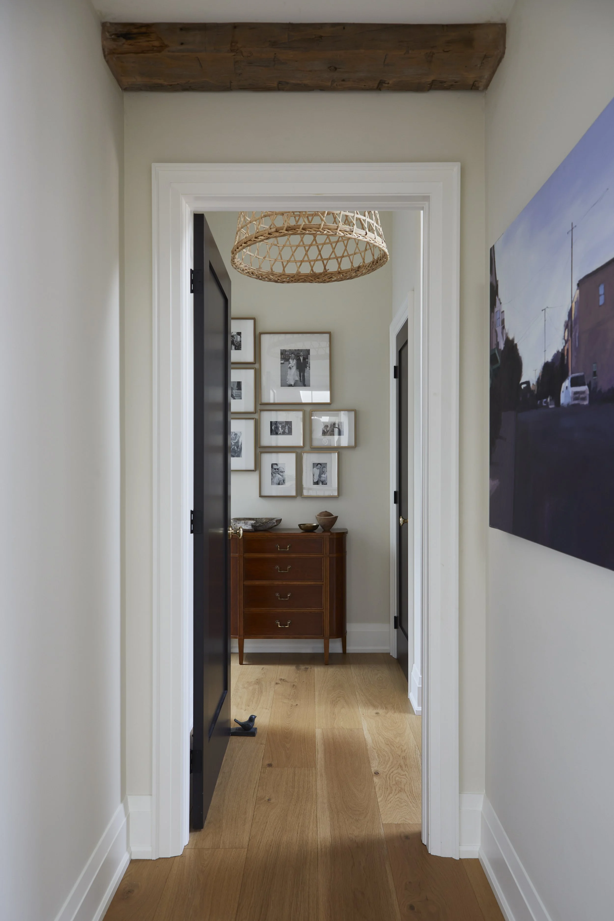 A view of a hallway leading to a small room with a wooden dresser, framed black-and-white photographs on the wall, and decorative bowls on top. The hallway has white walls, wooden flooring, and a hanging woven light fixture.