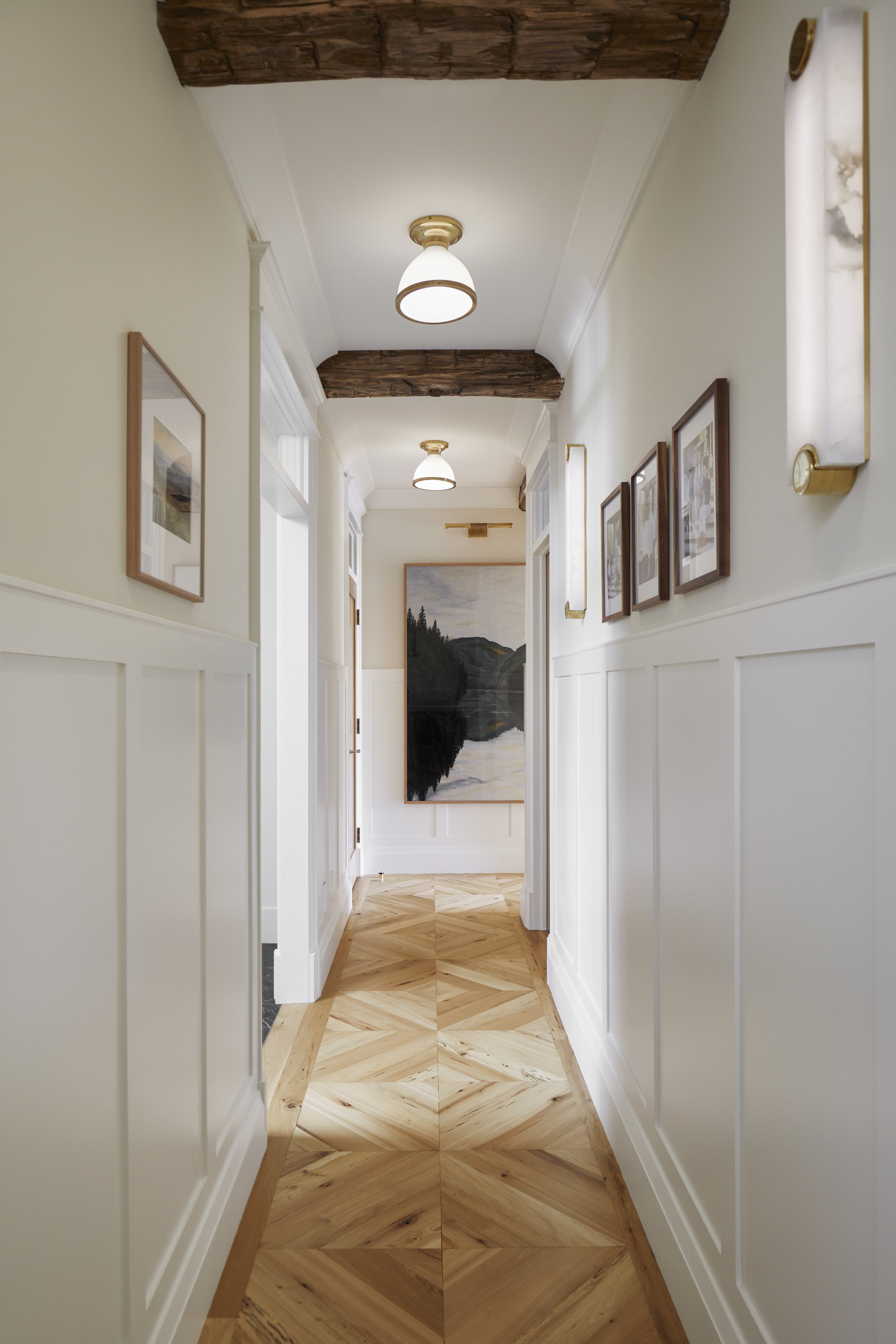 A hallway with wooden parquet flooring, white paneled walls, and framed artwork. The ceiling has two antique-style light fixtures and exposed wooden beams.