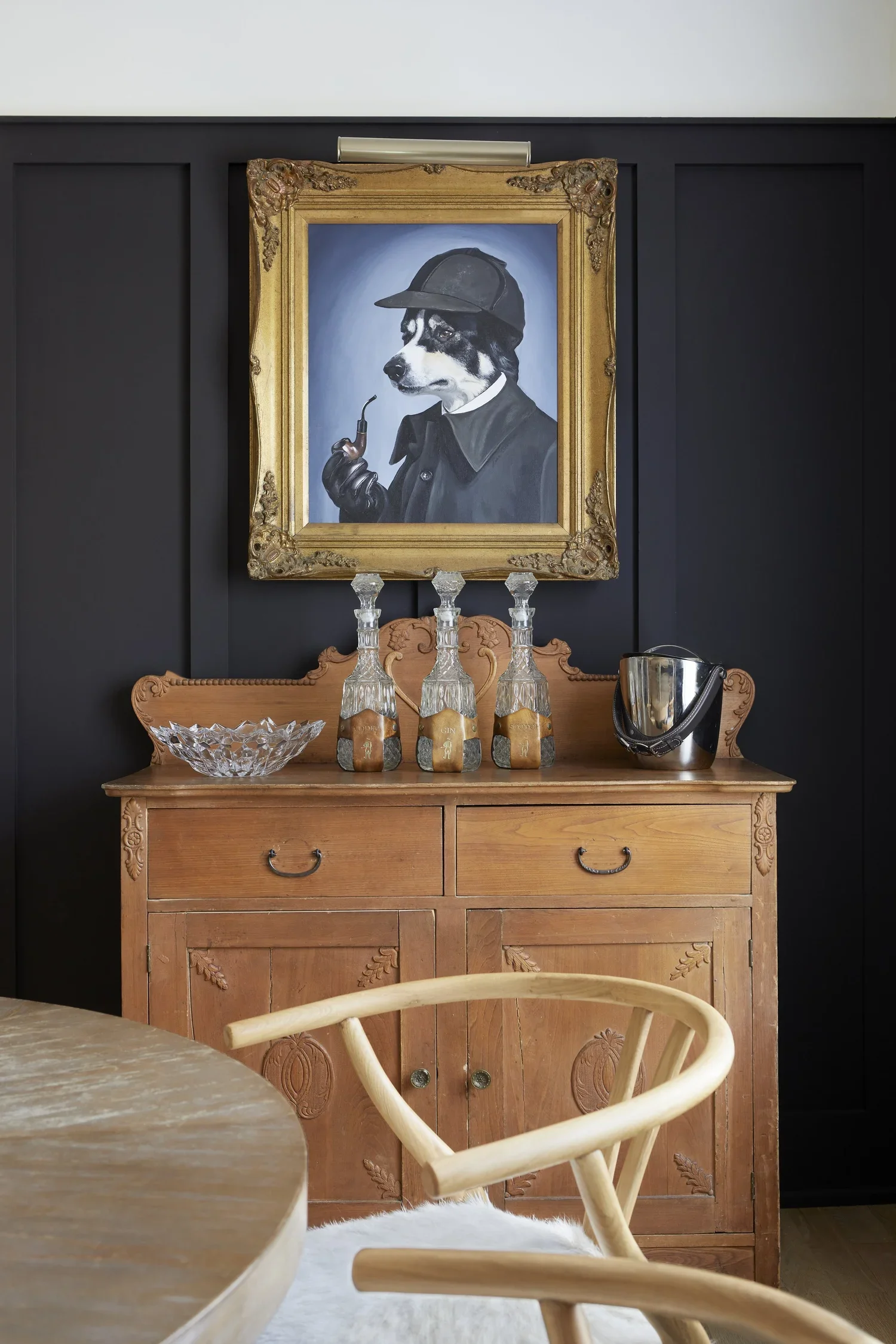 A wooden sideboard with three bottles, a glass bowl, and a party bucket on top. Above it, a framed portrait of a dog dressed as Sherlock Holmes with a hat, pipe, and coat, set against a dark wall.