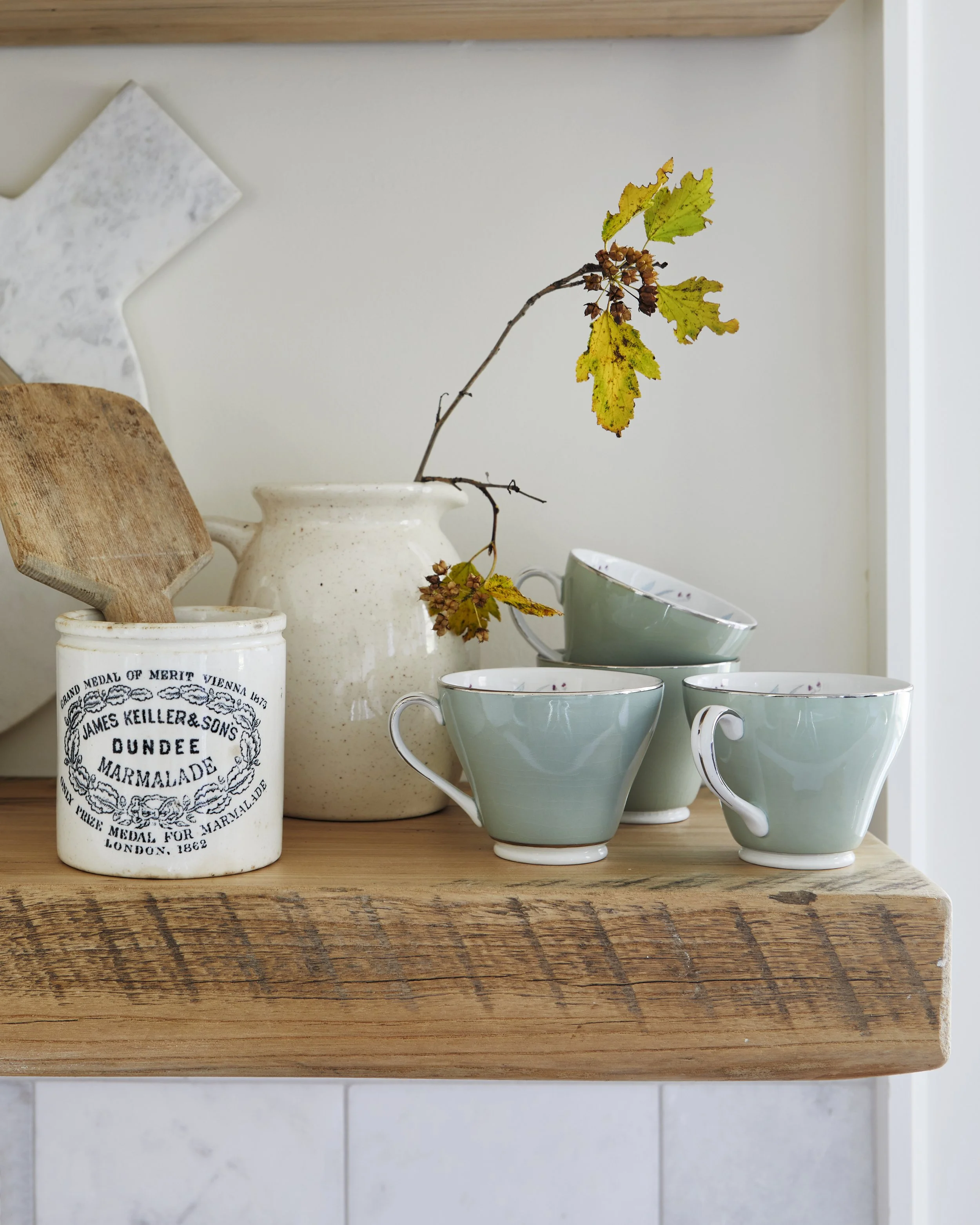 Ceramic cups stacked on a wooden shelf with a decorative jar containing a wooden spatula, a ceramic pitcher with a branch and leaves, against a light-colored wall.
