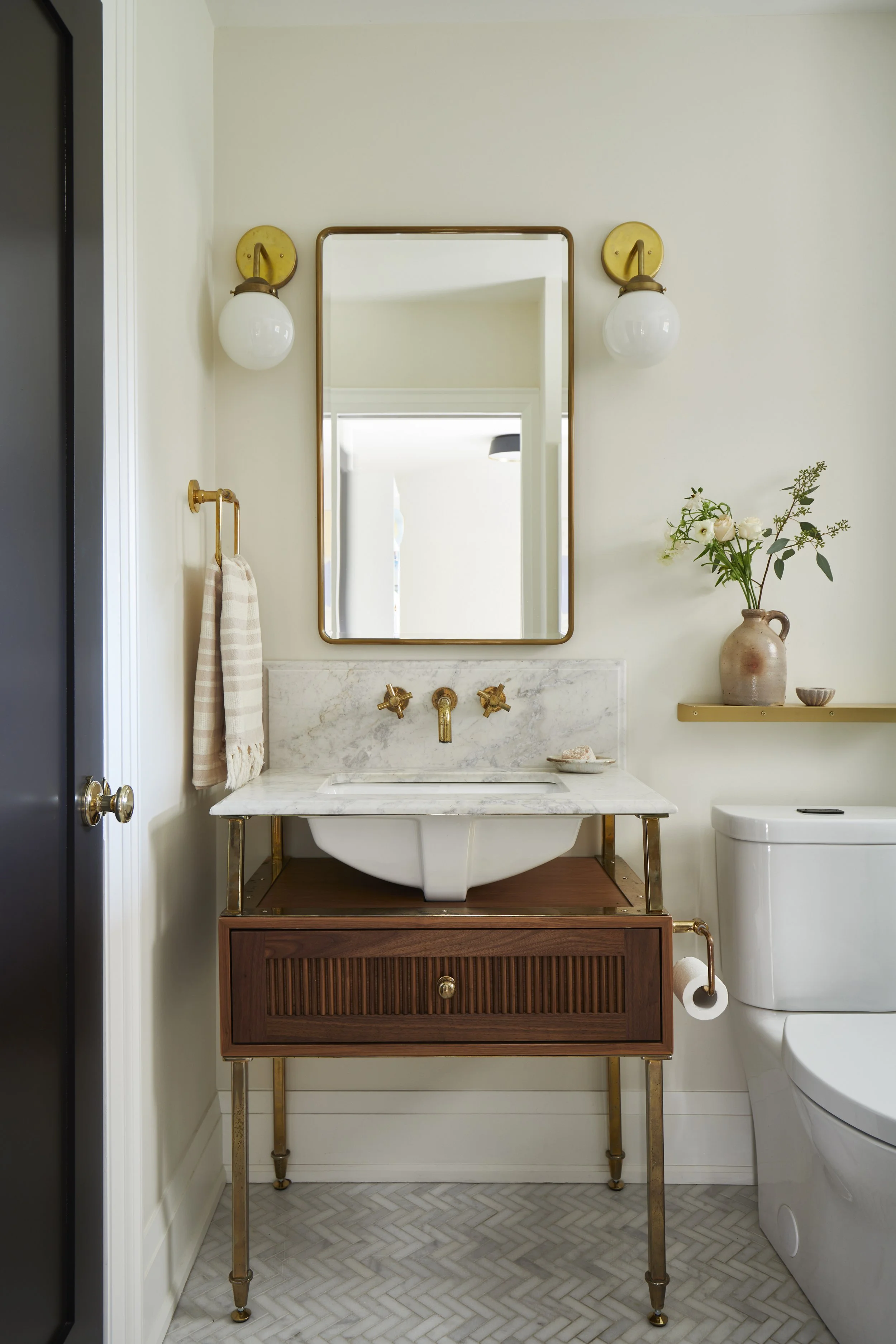 Bathroom with a vintage wooden vanity holding a vessel sink, gold fixtures, marble countertop, wall-mounted mirror, wall sconces, a small vase with flowers, and a toilet on the right.