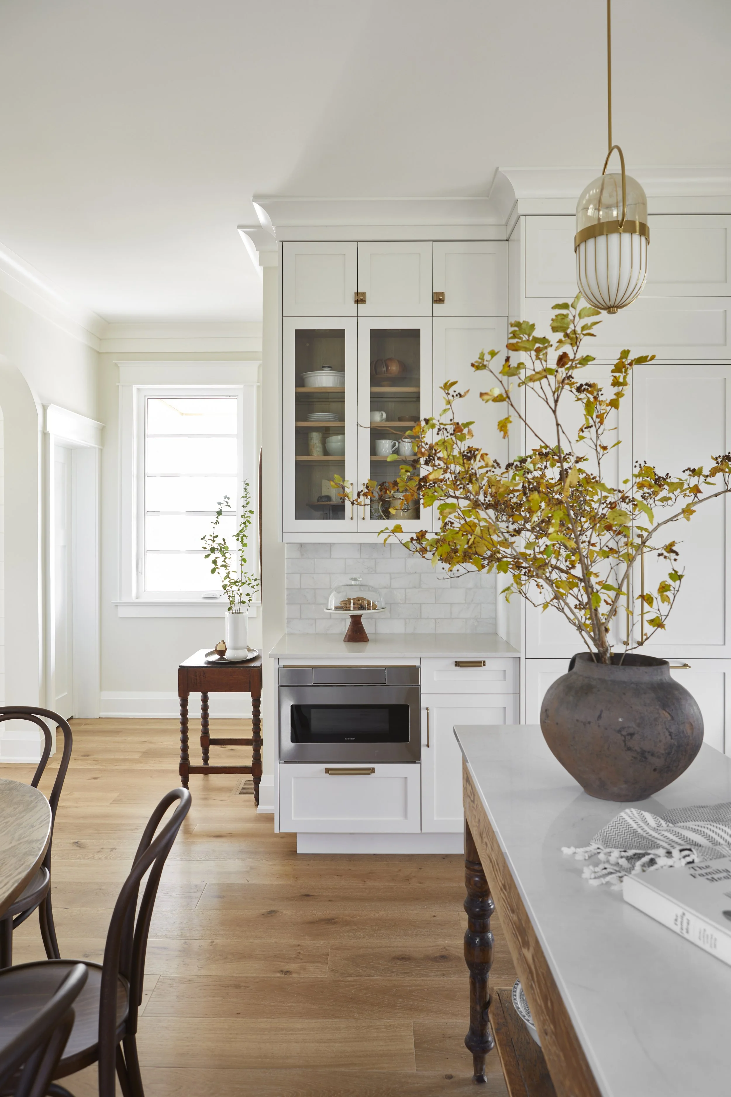 A bright, modern kitchen with white cabinets, hardwood floors, a large window, and a countertop with a large vase containing greenery.