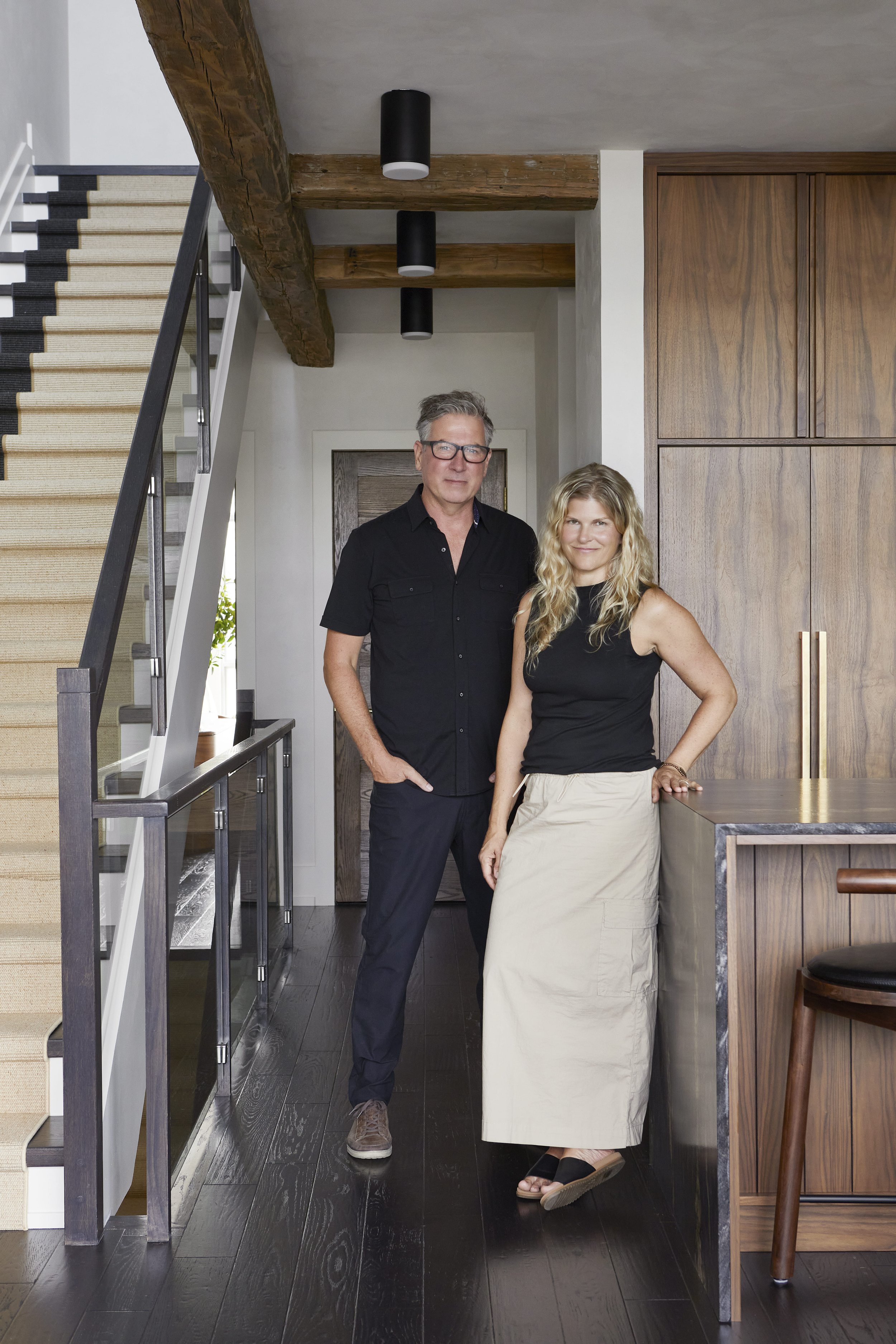 A man and woman stand together inside a modern home, near a wooden kitchen island, with a staircase on the left and a wooden ceiling beam overhead.