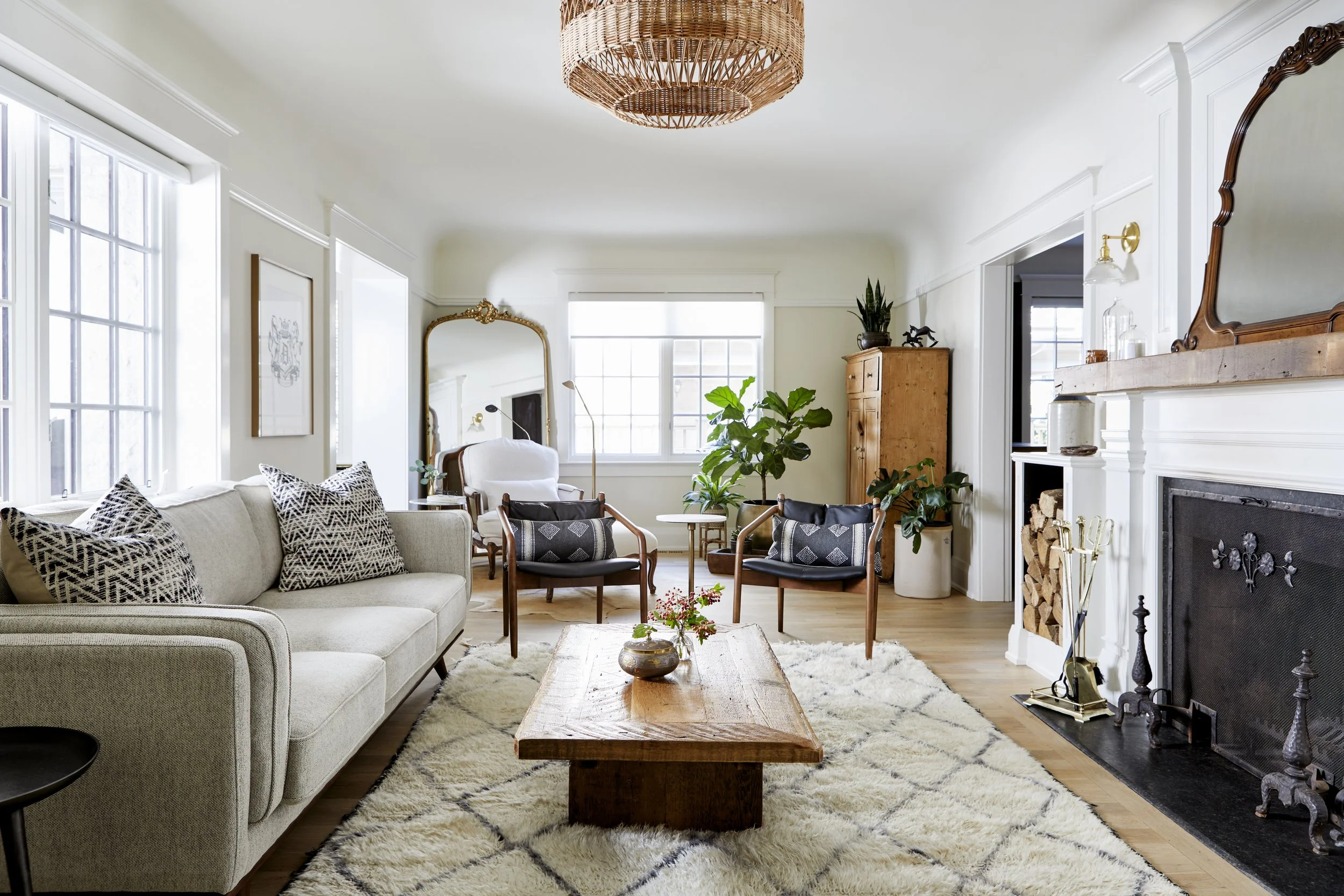Bright living room with white walls, large windows, a beige sofa with black and white patterned pillows, a wooden coffee table, black armchairs, a white armchair, a large green houseplant, and a fireplace with firewood stacked inside. Decor includes 