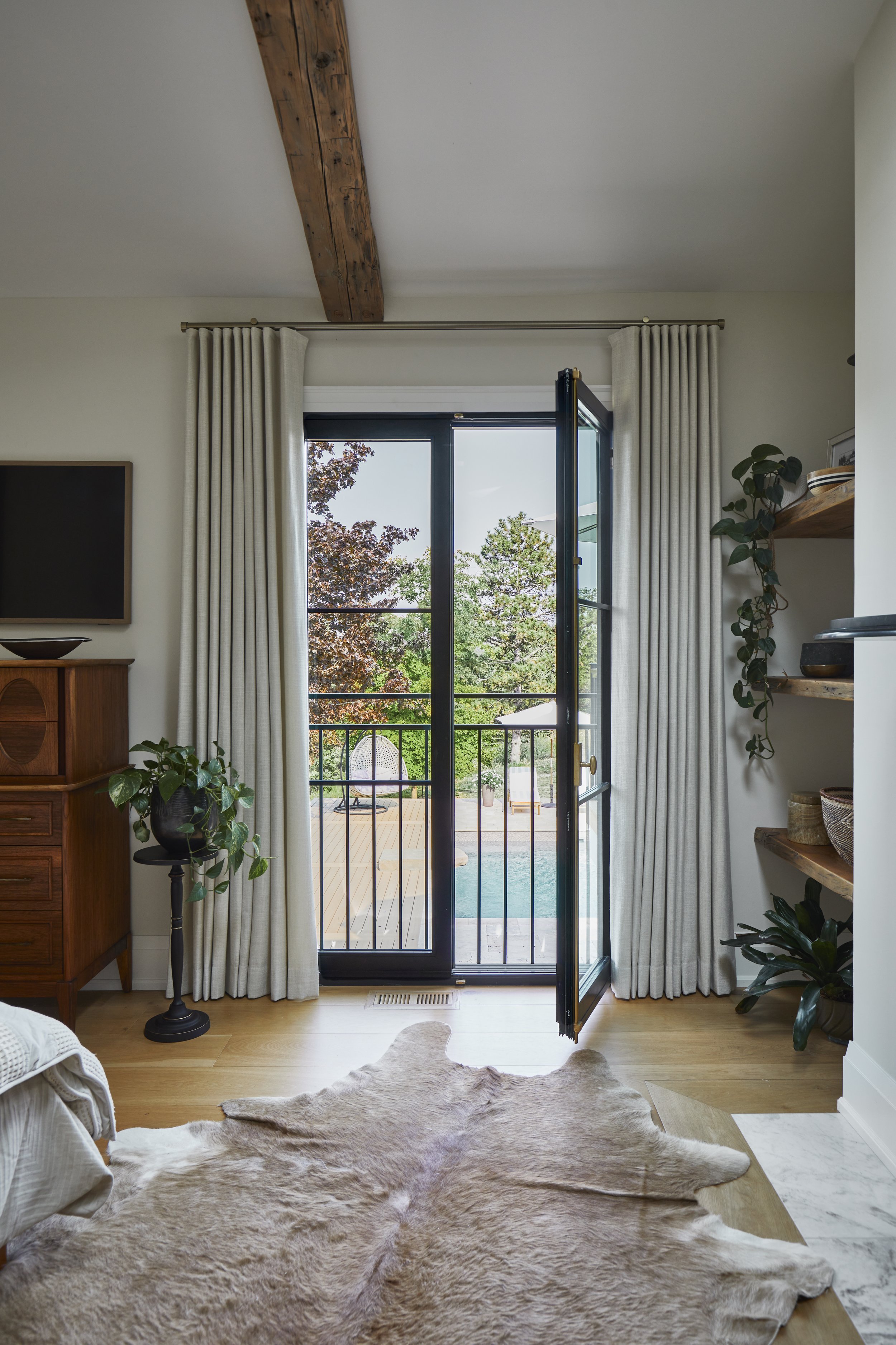 A bedroom with an open door leading to a wooden deck with a view of trees and a pool outside. The room has light-colored curtains, wooden shelves, and plants, with a cowhide rug on the floor.