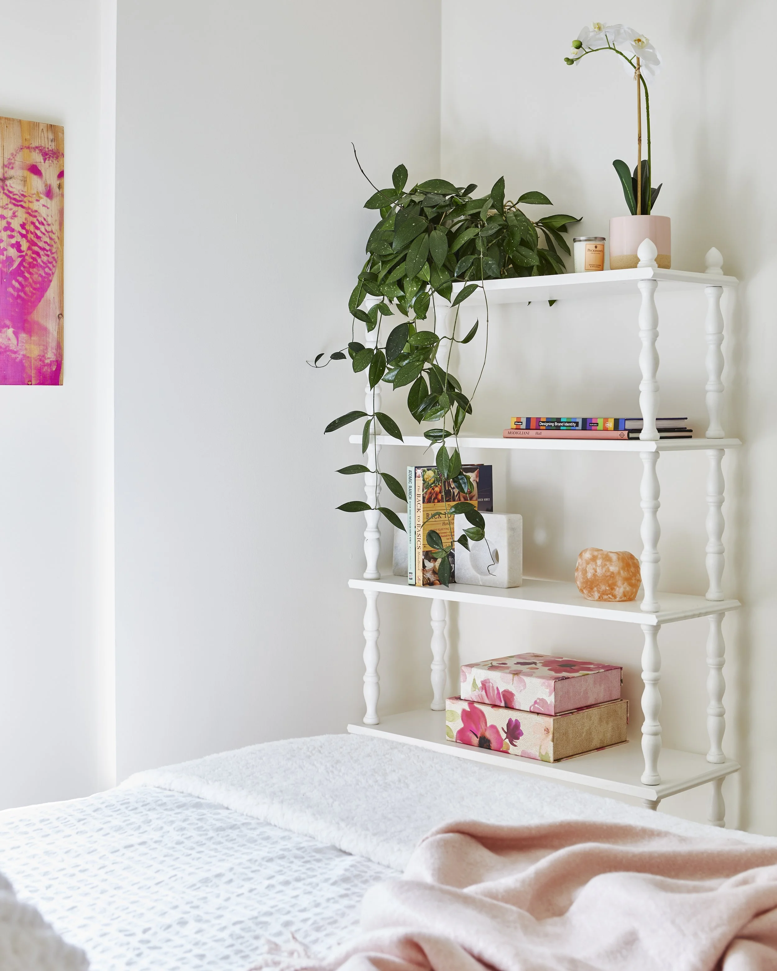 A white bookshelf with potted plant, books, salt lamp, and decorative boxes, next to a bed with pink blanket and white bedding.