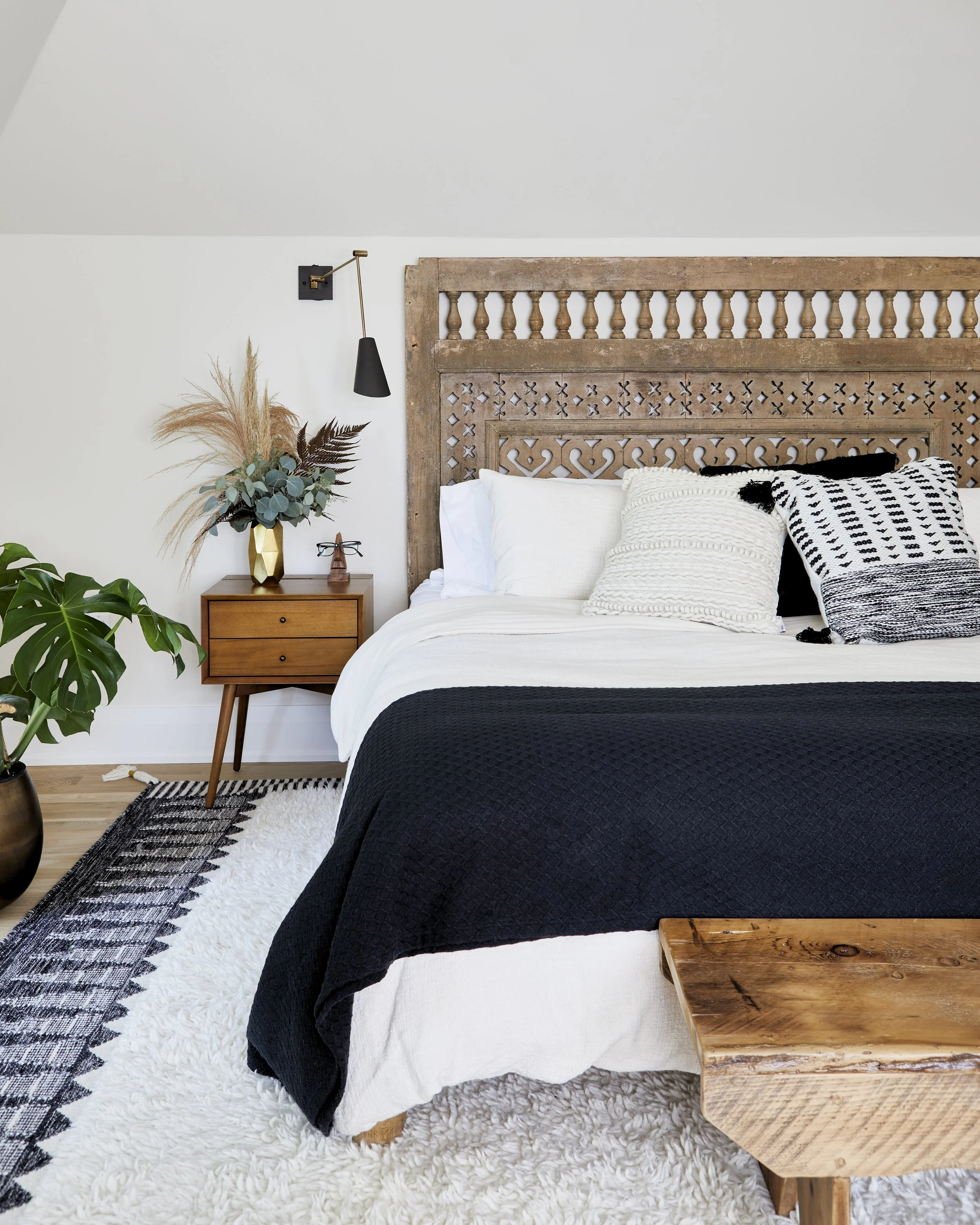 A cozy bedroom with a wooden headboard, white bed linens, black and white pillows, a black blanket, a wooden bedside table with a vase of dried flowers, a plant in a black pot, and a black and white rug.