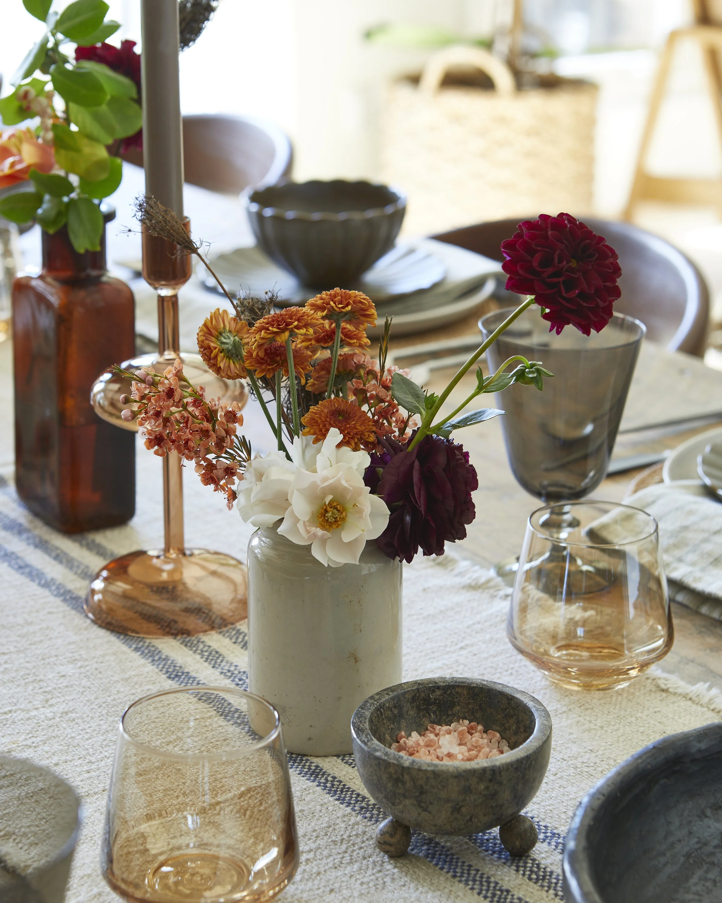 A dining table decorated with a white vase of colorful flowers, pink salt in a stone bowl, amber-colored glassware, and various dishes and napkins.