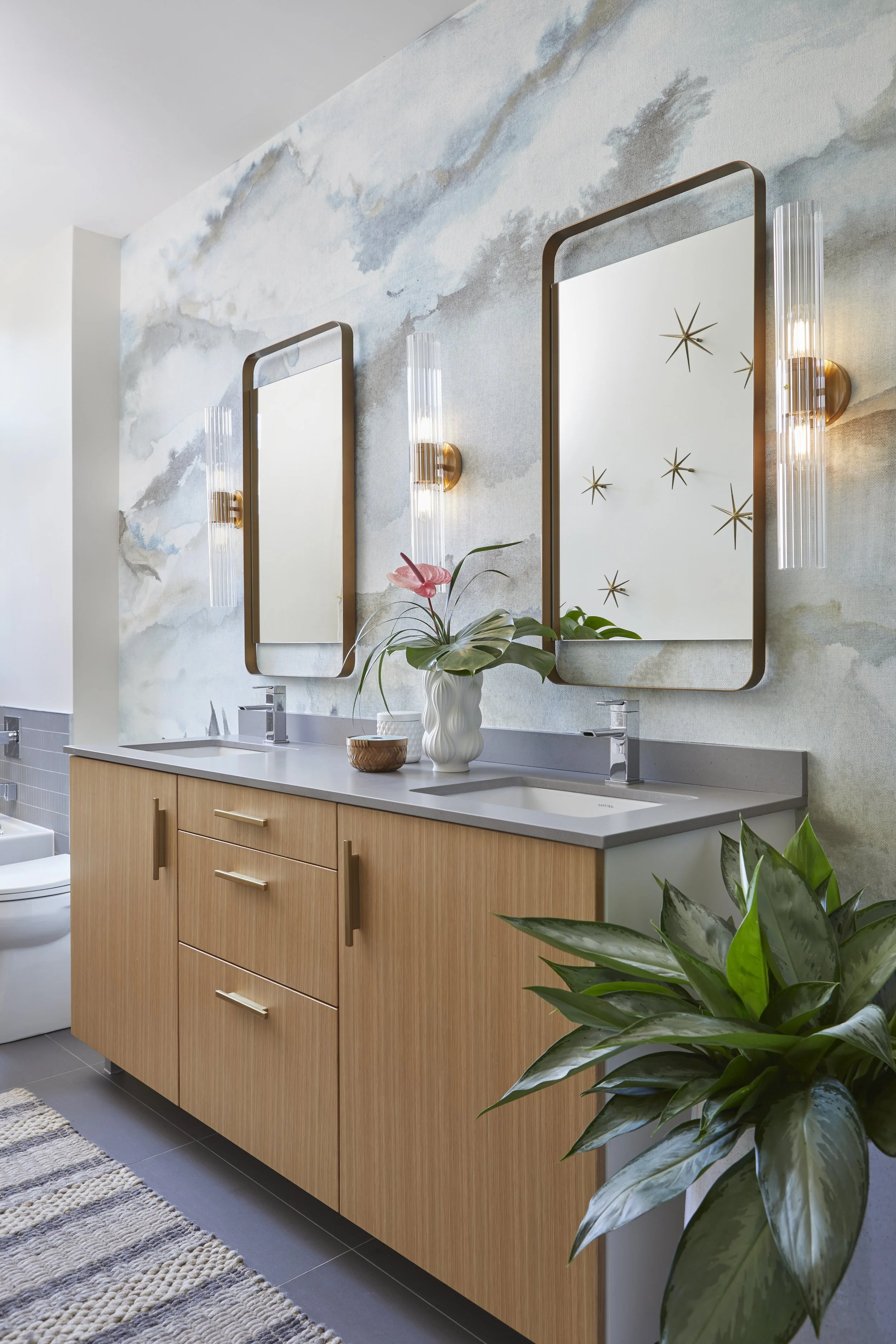 A modern bathroom with double sinks, rectangular mirrors, wall-mounted light fixtures, and wooden cabinet storage. The wall features a subtle watercolor pattern with gold starburst decals, and there are green plants on the countertop and in front of 
