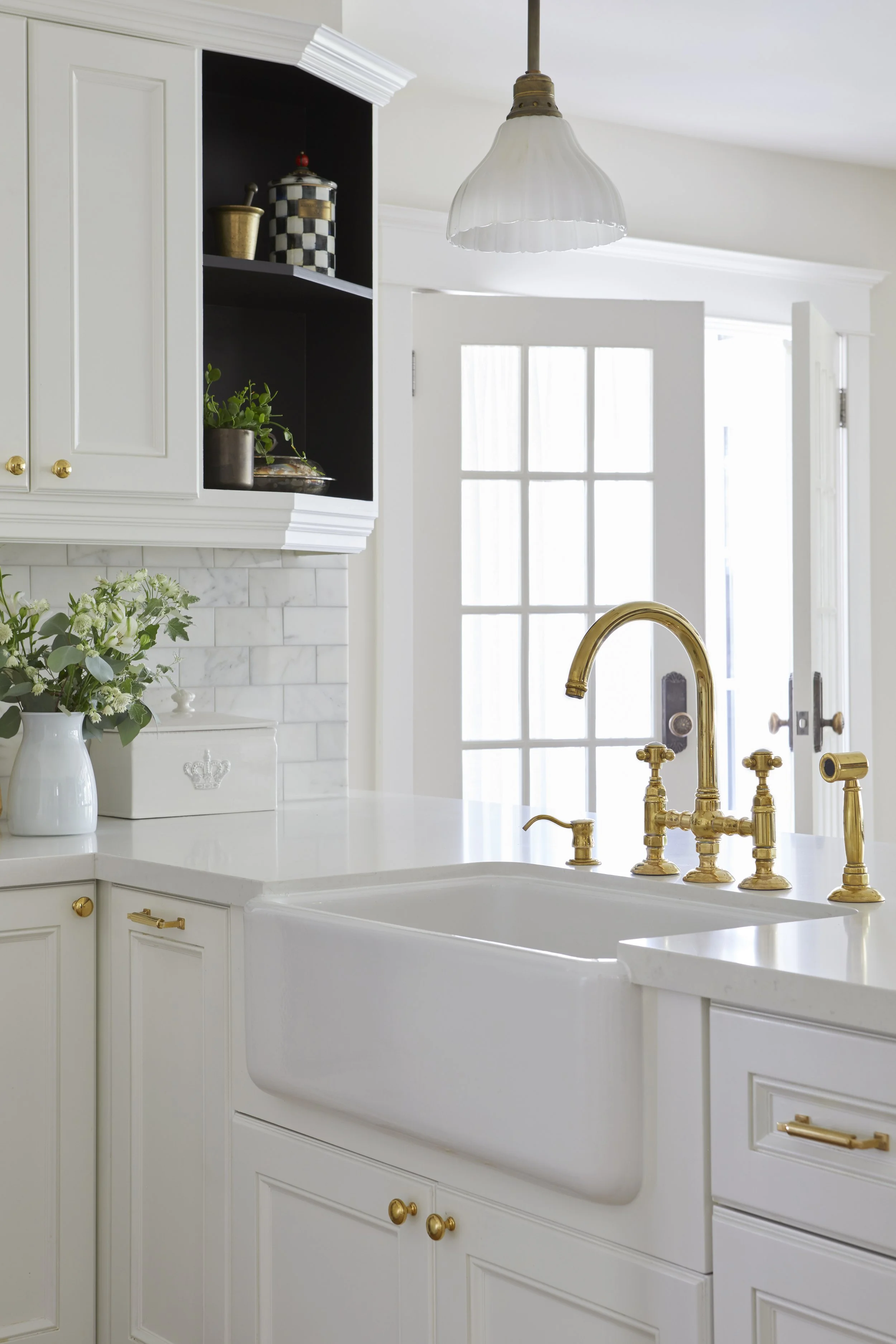 White kitchen with gold fixtures, a farmhouse sink, a window with sunlight, and decorative items on the shelves.