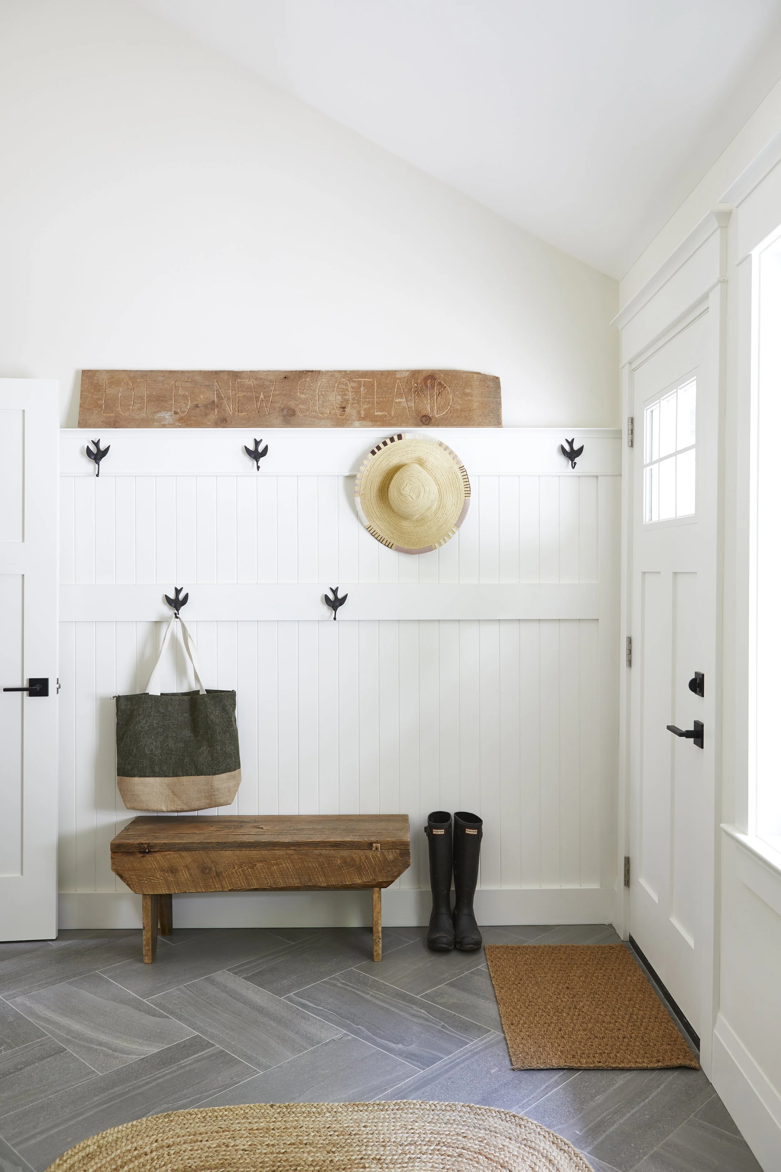 Entryway with white paneled walls, a wooden bench, black rubber boots, a hanging tote bag, decorative black hooks, a straw hat, and a wooden sign that reads 'Lot 45 New Scotland'