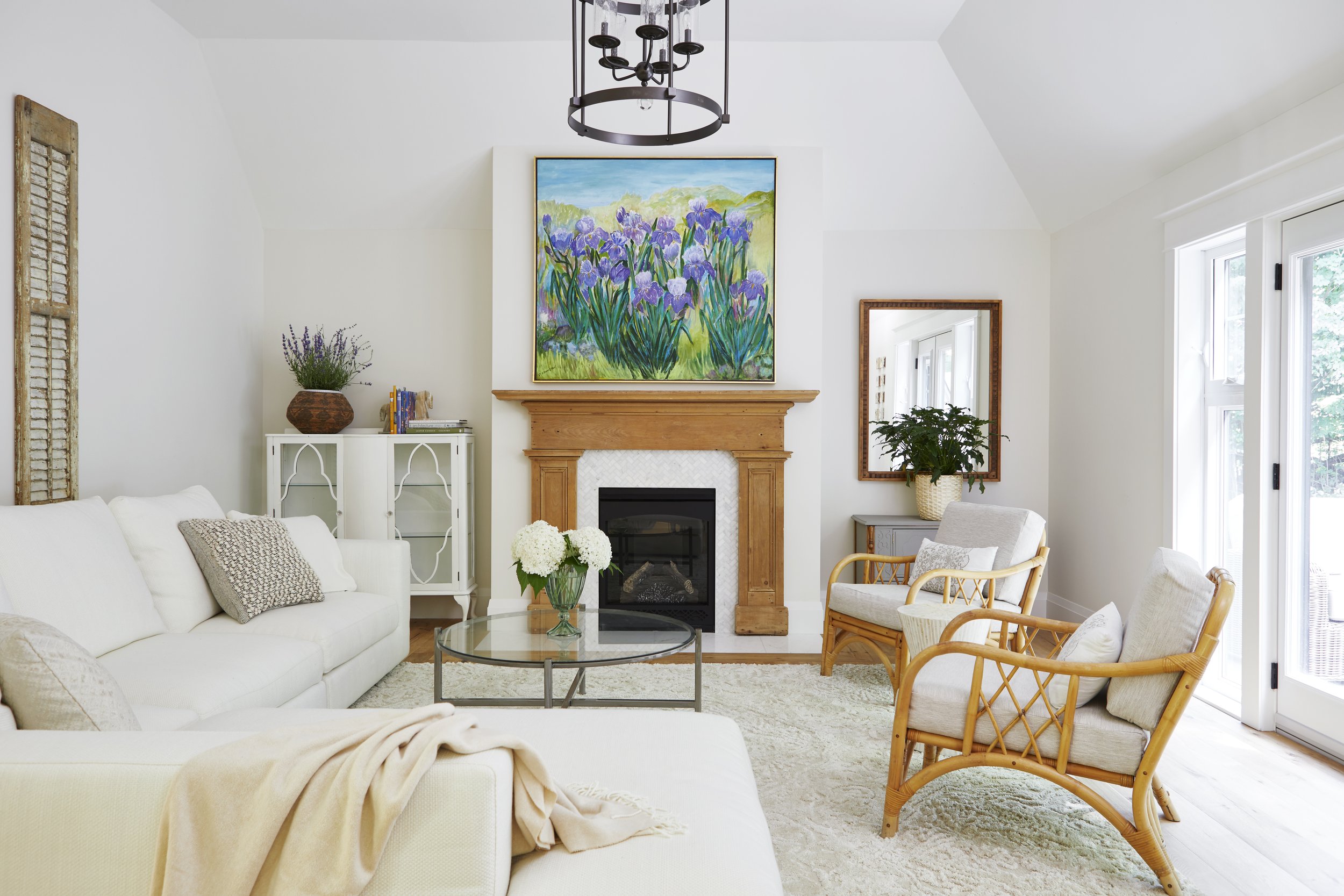 Bright living room with white walls, a fireplace with a wooden mantel, a colorful floral painting above, a beige sofa with cushions, rattan chairs, a glass coffee table with white flowers, and large windows with sliding glass doors.