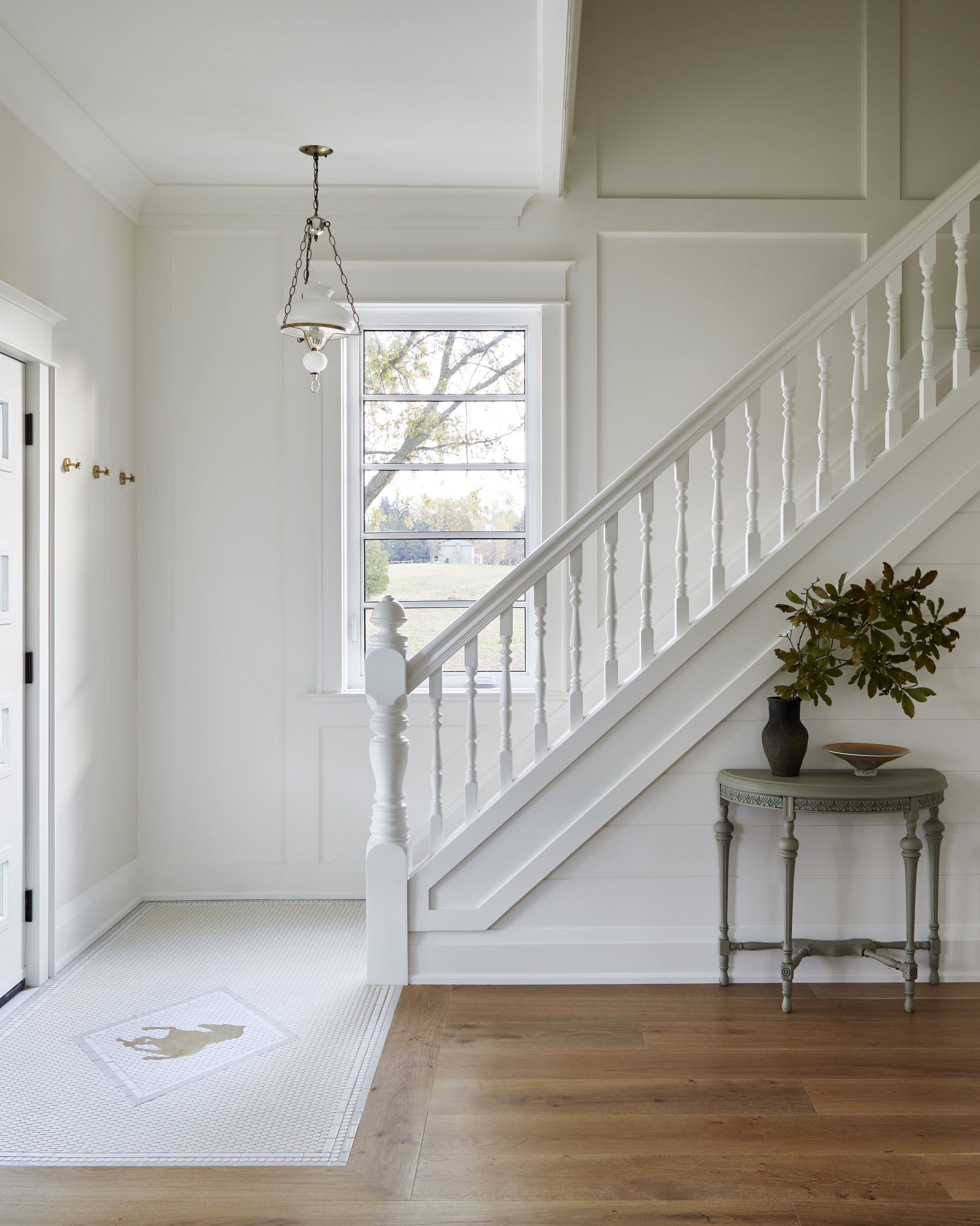 Interior view of a bright, white entryway with a staircase, large window, and wooden flooring. A small table with a vase and plant is under the stairs, and a ceiling light fixture hangs from the ceiling.