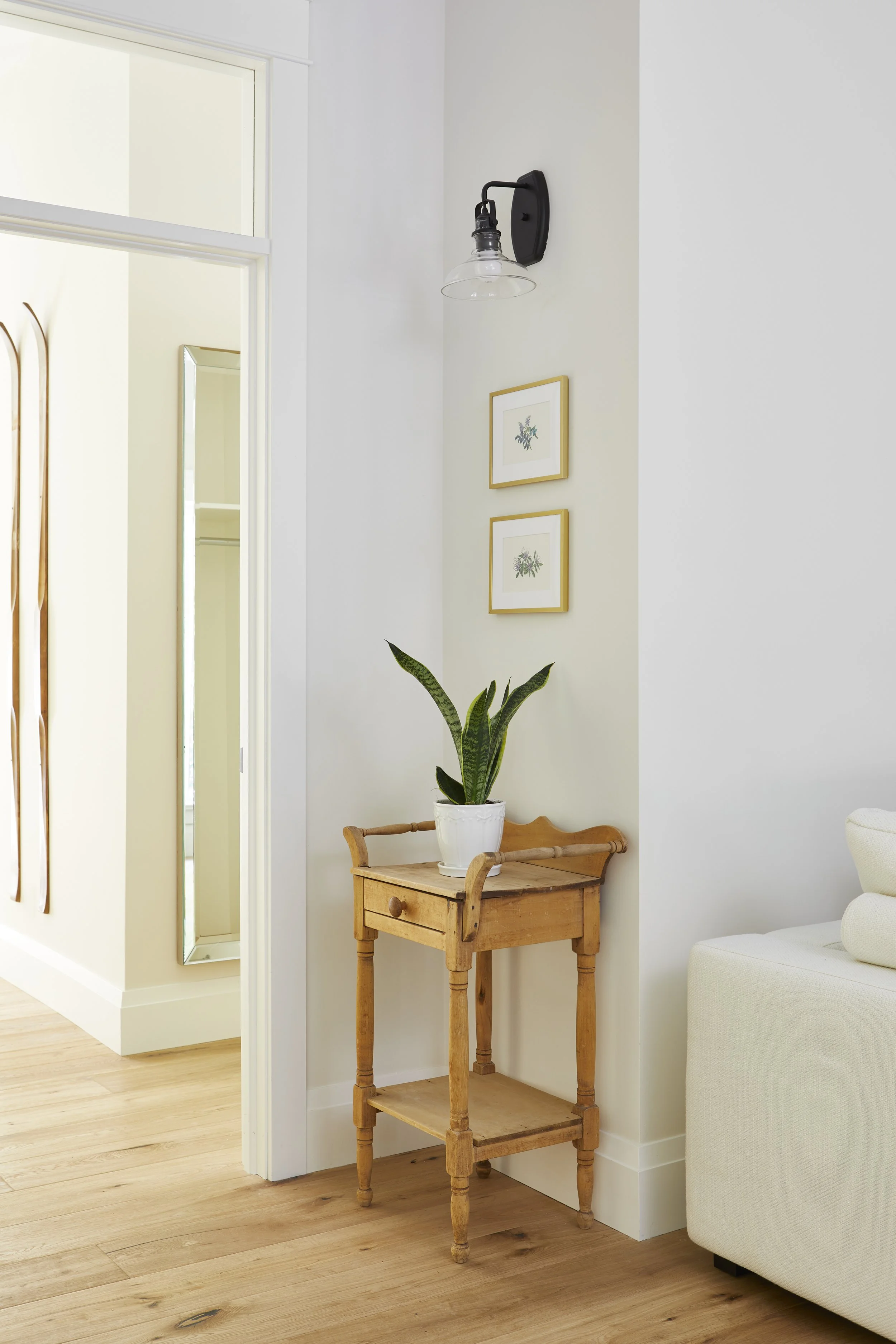 A corner of a room with a wooden side table holding a potted snake plant, framed botanical prints on the wall, a wall-mounted black sconce with a clear glass shade, a partially visible white sofa, white walls, and light-colored hardwood flooring.
