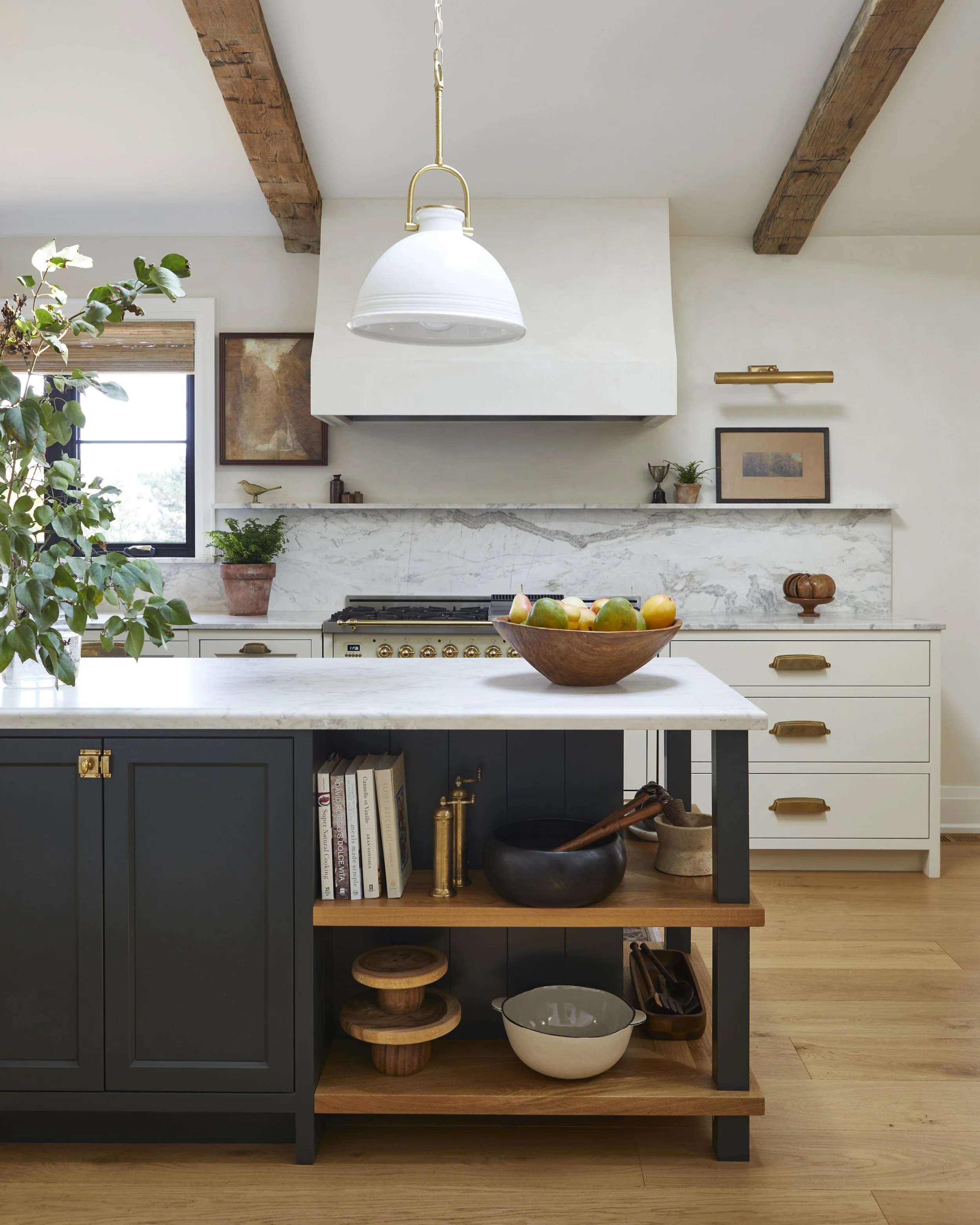 A modern kitchen with a white marble countertop, white and dark blue cabinets, wooden shelves, and exposed wooden beams on the ceiling. There is a bowl of fruit on the island, potted plants, framed artwork, and decorative objects.
