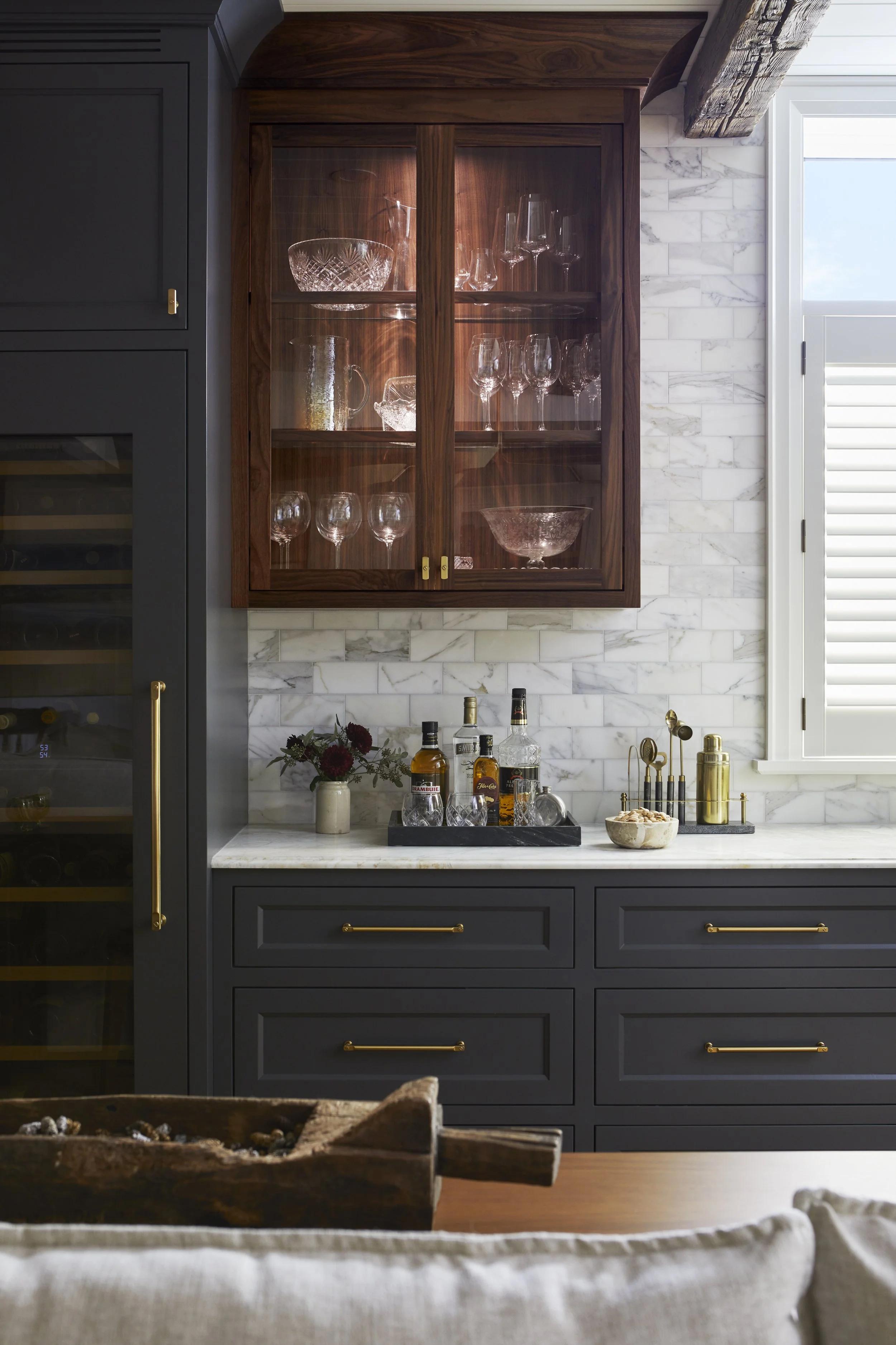 Interior view of a modern bar area with dark gray cabinets, gold handles, and a marble countertop. A wooden glass cabinet displays glassware, and a window with shutters lets in natural light. The countertop has liquor bottles, a bowl, and decorative 