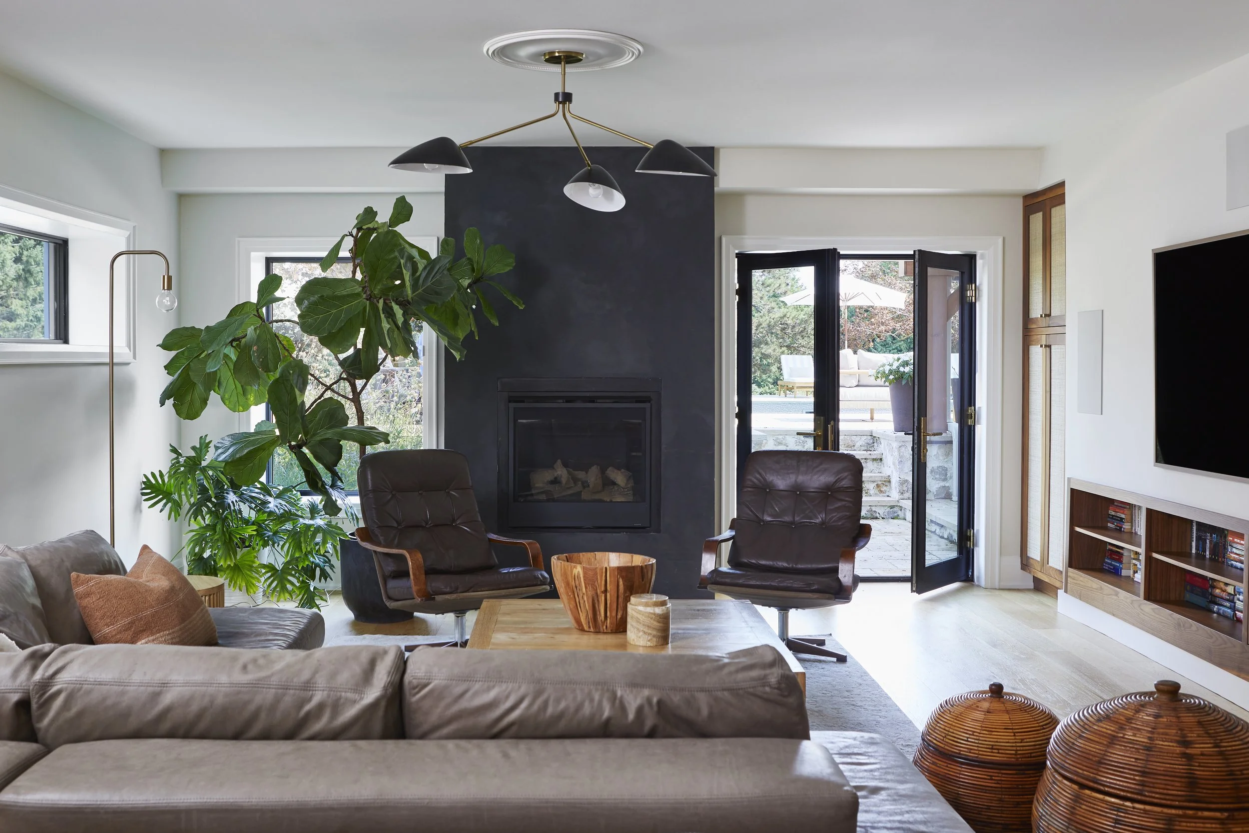 Modern living room with a black fireplace, brown leather chairs, a beige sofa, wooden coffee table, large green plants, and a door leading outside to a patio with outdoor furniture.