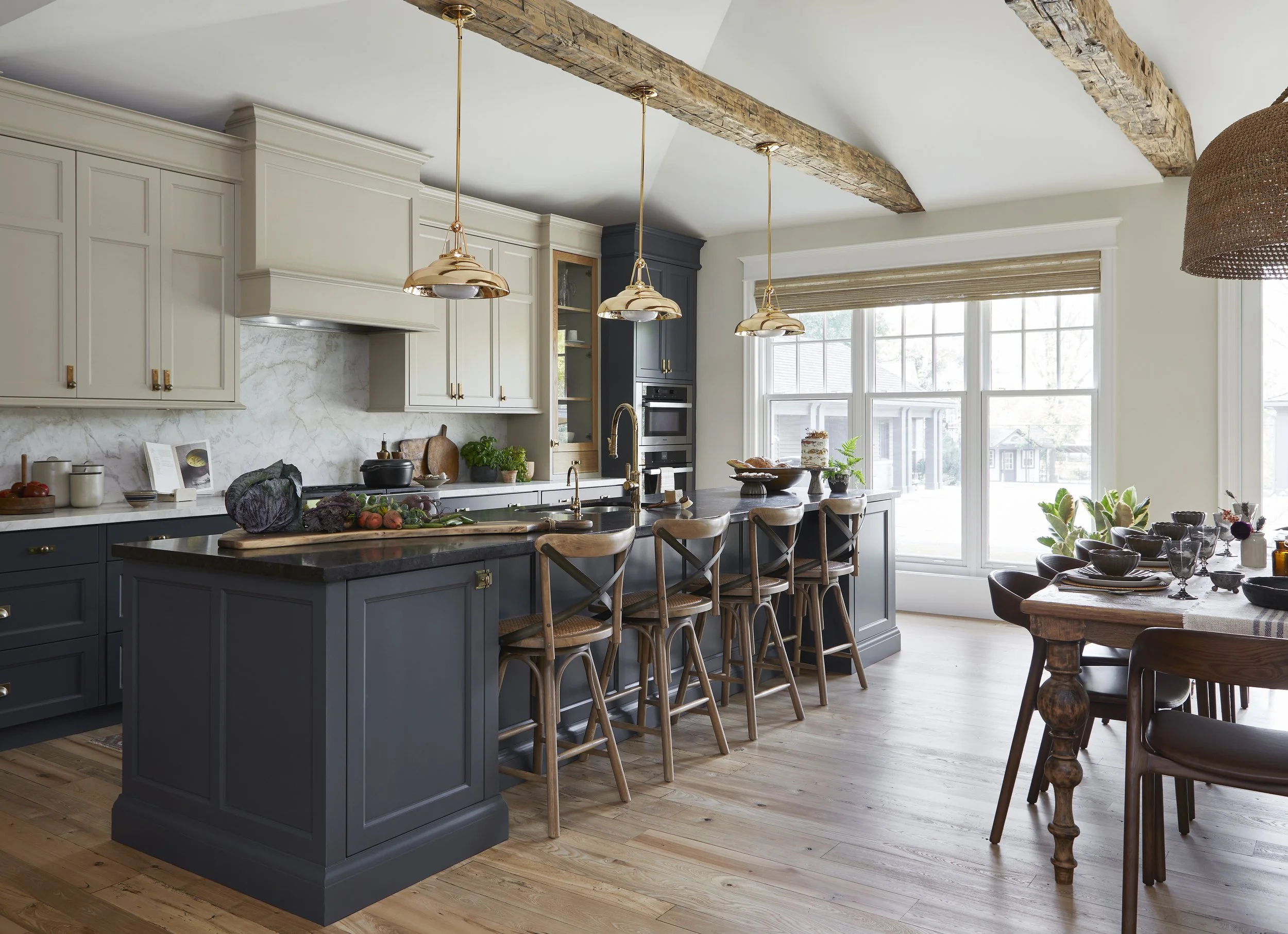 Modern kitchen with white and blue cabinetry, marble backsplash, and large window. Features wooden flooring, pendant lighting, and countertops with various decor and vegetables.