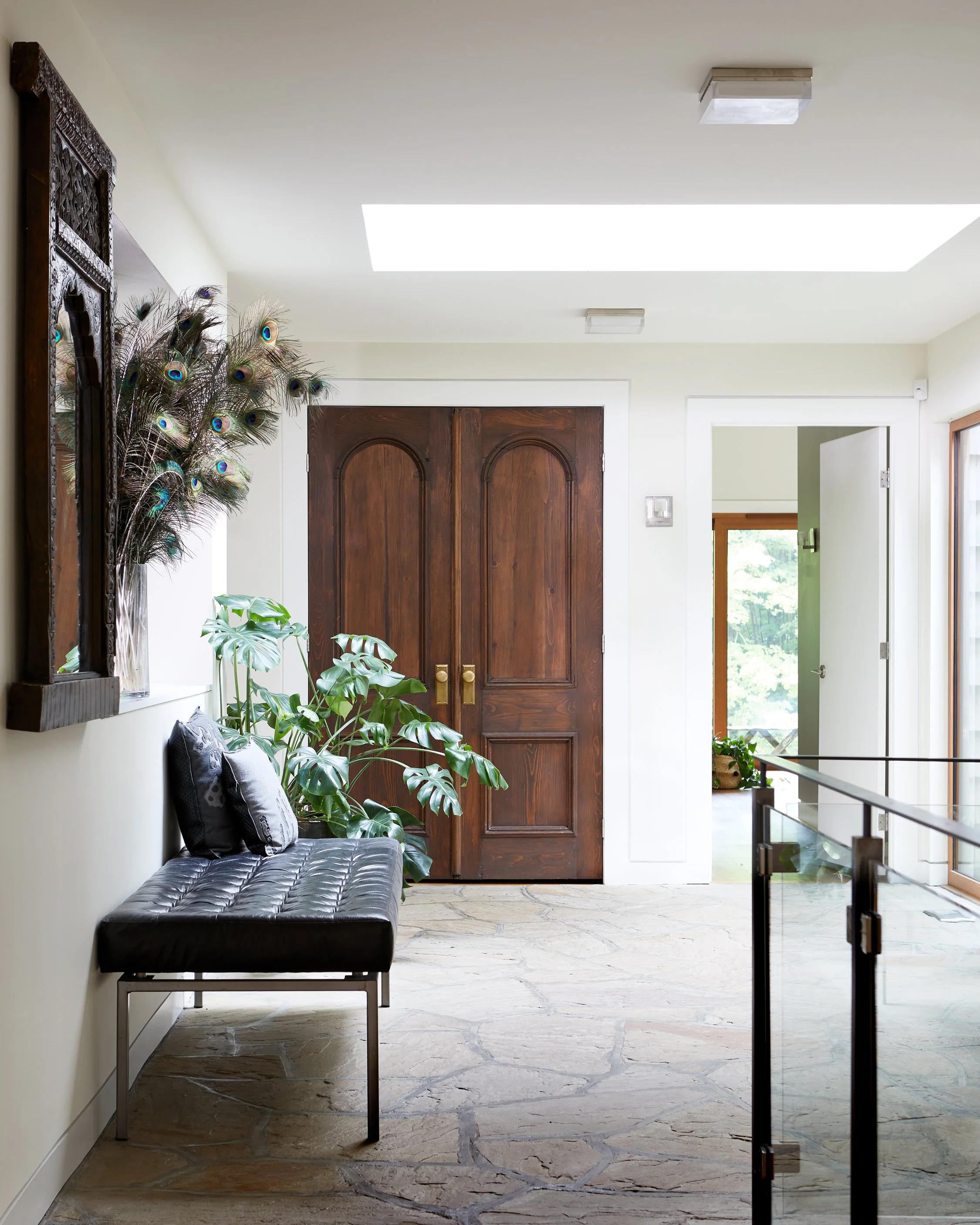 Entryway with wooden double doors, a black leather bench, and a large green plant, with peacock feathers decor on the wall.