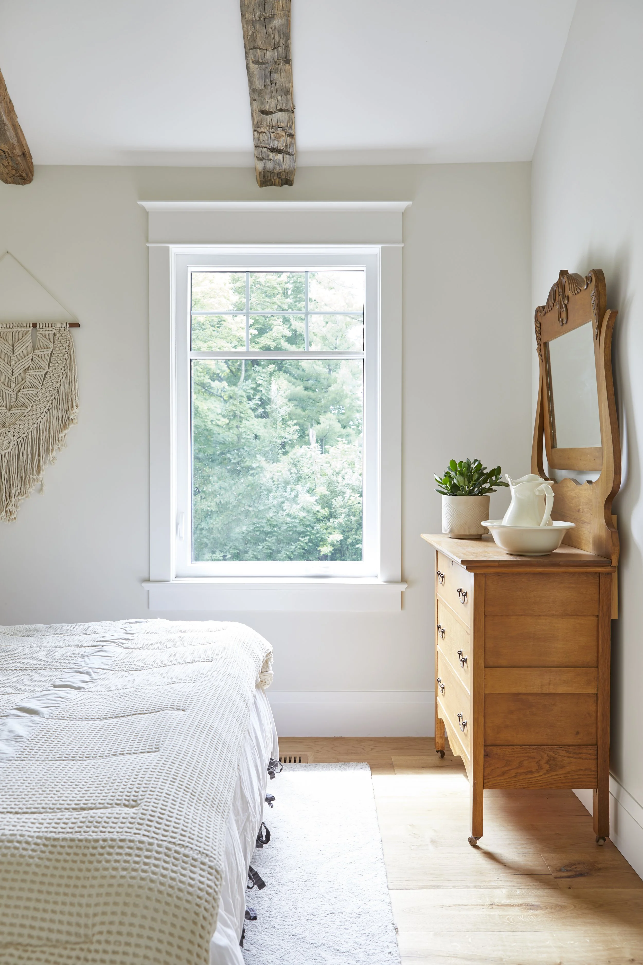 A bedroom corner with a bed, a window showing green trees outside, and a wooden dresser with a mirror, plant, pitcher, and bowl.