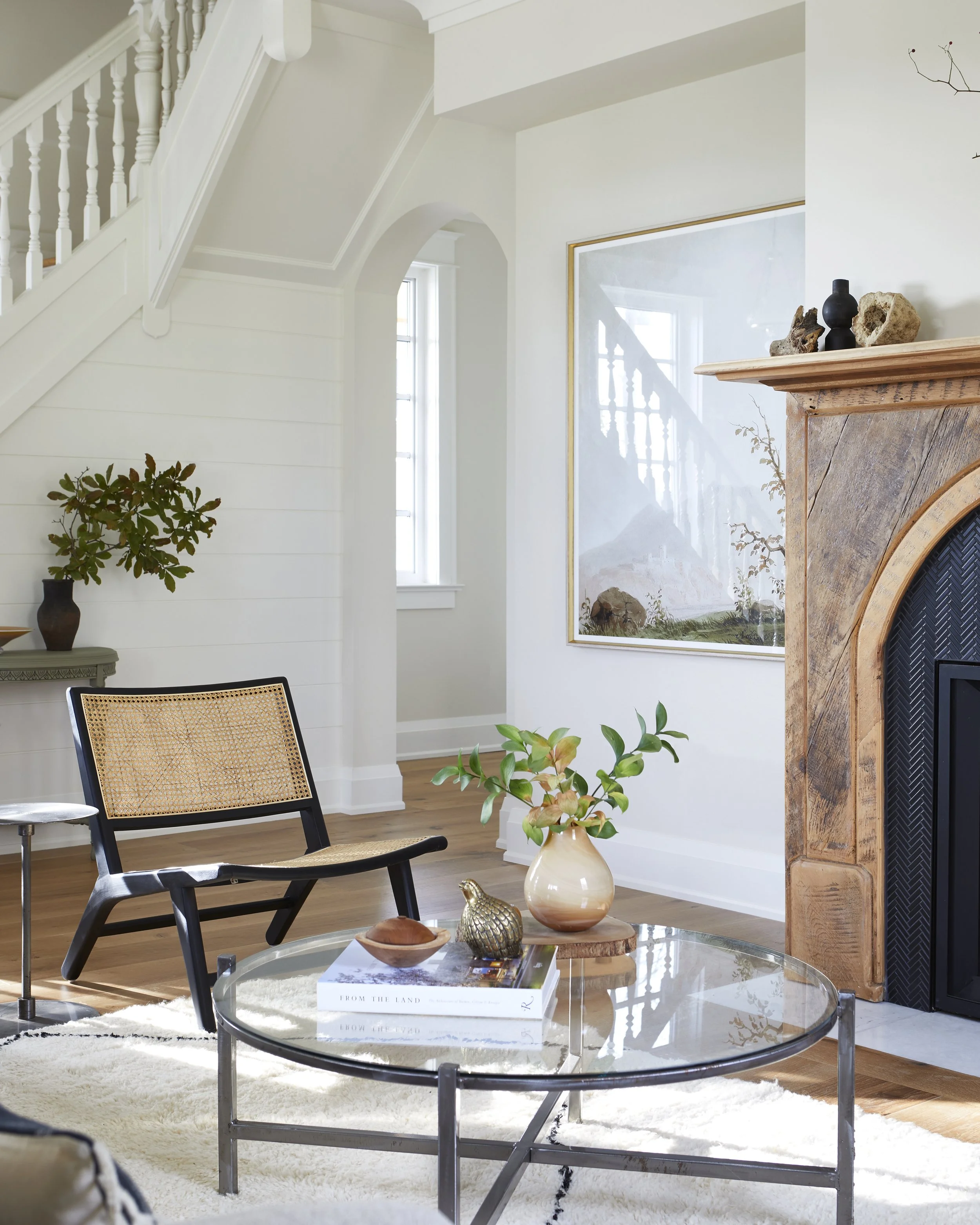 Living room with white walls, a wooden fireplace, a glass-top coffee table, and a black chair with woven rattan backrest. Decor includes plants, a large mirror, and decorative stones on the mantel.