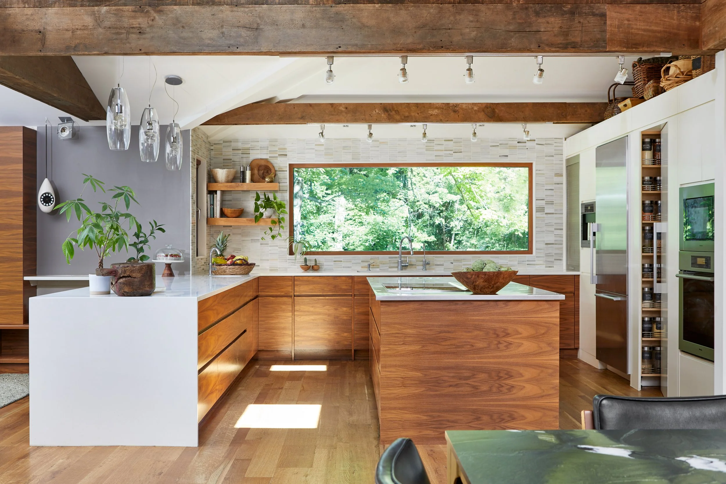 Modern kitchen with wooden cabinets, a large window showing greenery outside, and a white kitchen island. There are plants, bowls, and glass pendant lights above.
