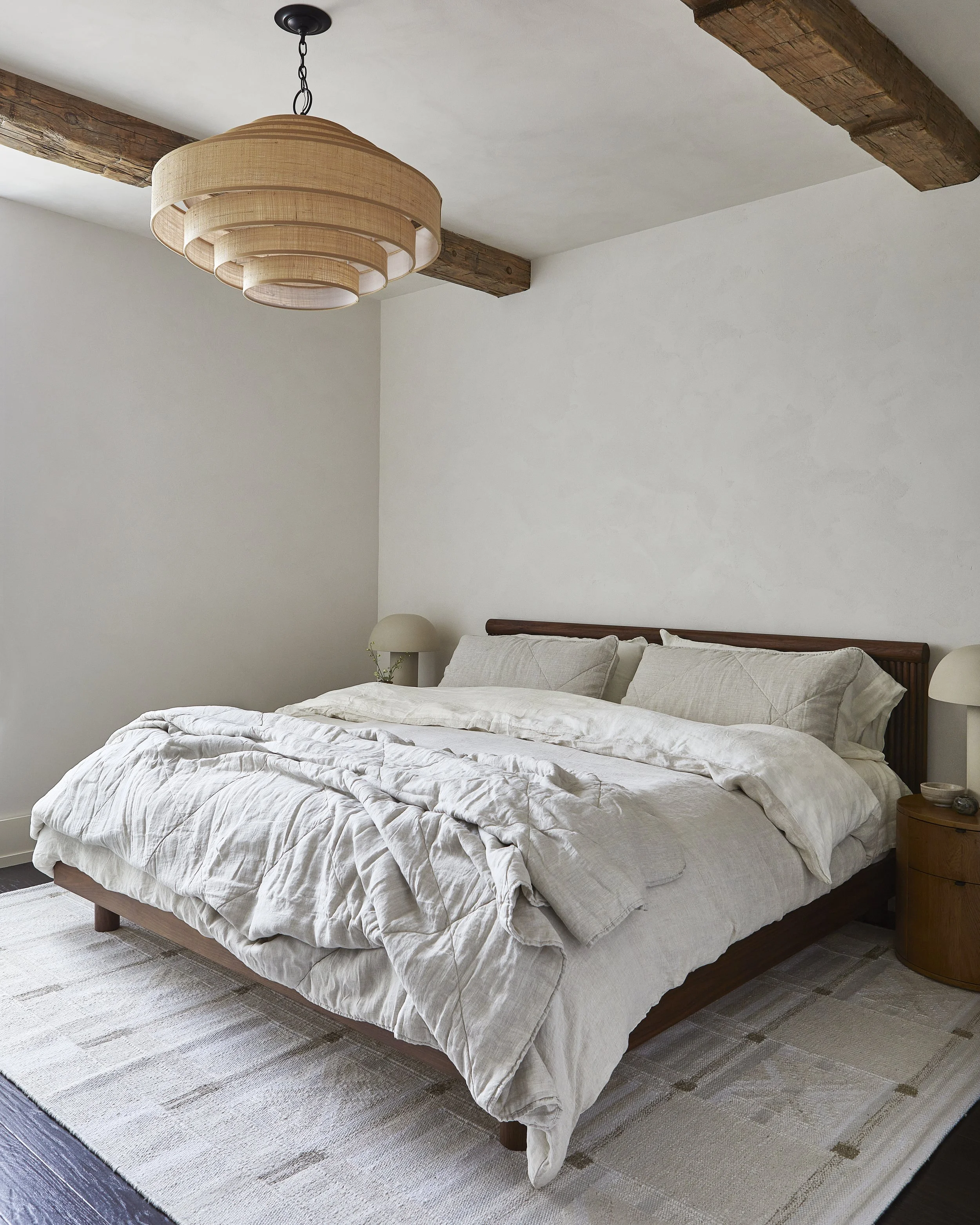 A neatly made bed with beige linens and pillows in a minimalist bedroom. There are two wooden bedside tables with small lamps, a textured beige carpet, white walls, and wooden beams on the ceiling. A modern layered pendant light hangs from the ceilin