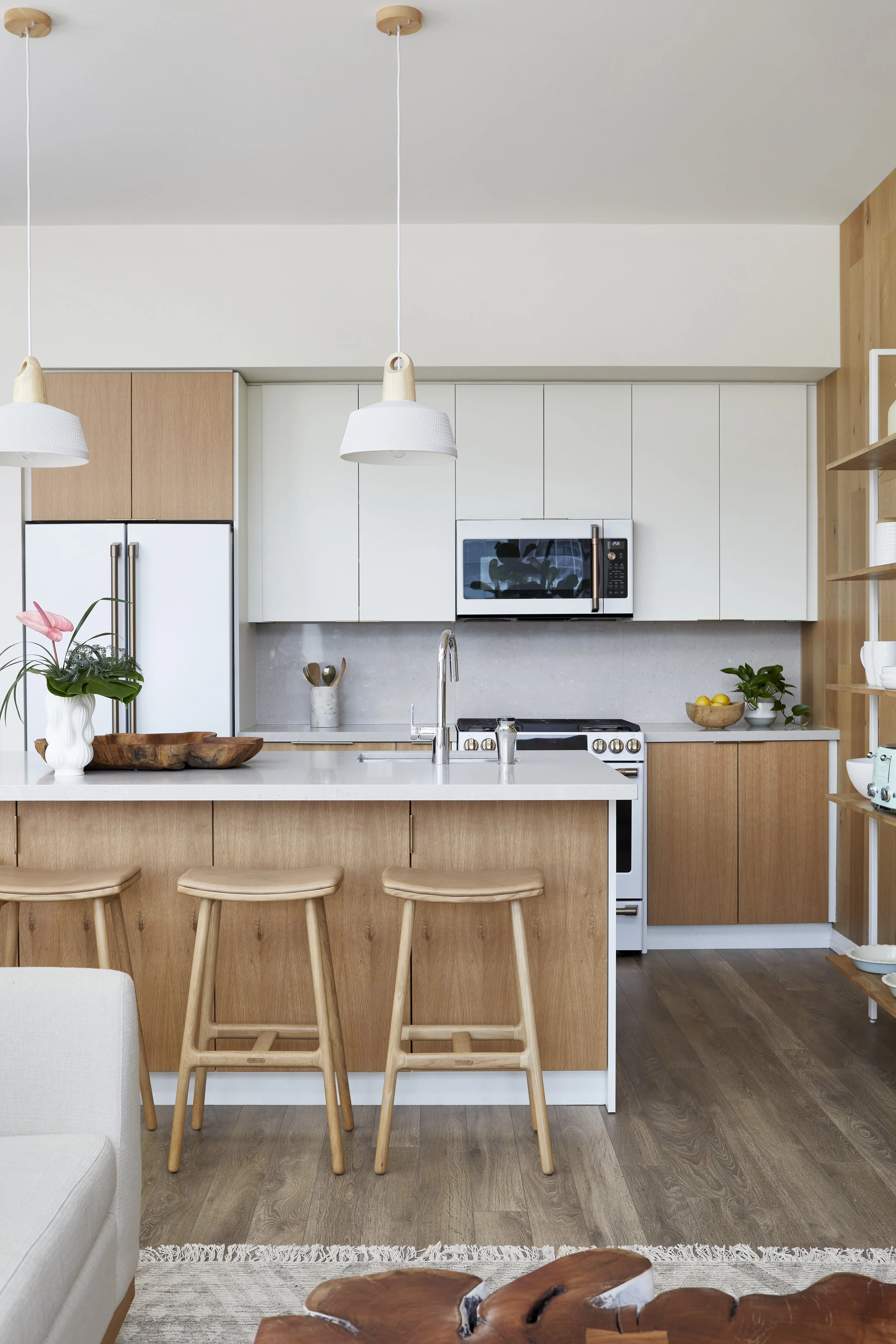 Modern kitchen with wooden cabinets, white countertops, and stainless steel appliances, including a microwave above the stove. There are three wooden bar stools at the counter, a vase with pink flowers, and a small plant with lemons on the side count