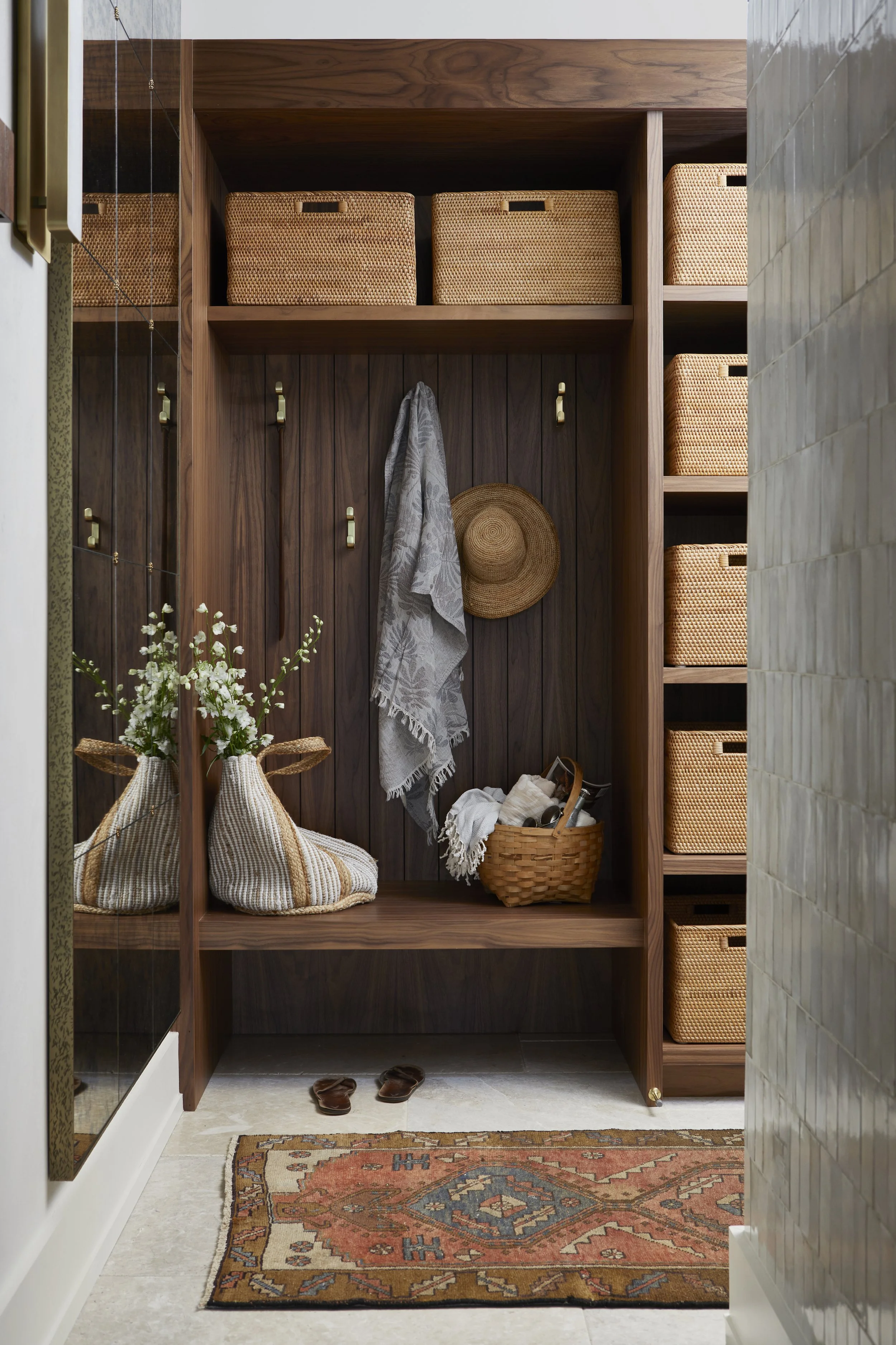 A cozy entryway with a wooden closet holding woven baskets, a hat and scarf hanging on hooks, a basket with shoes, a pair of sandals on a patterned rug, and a mirror reflecting a flower arrangement.