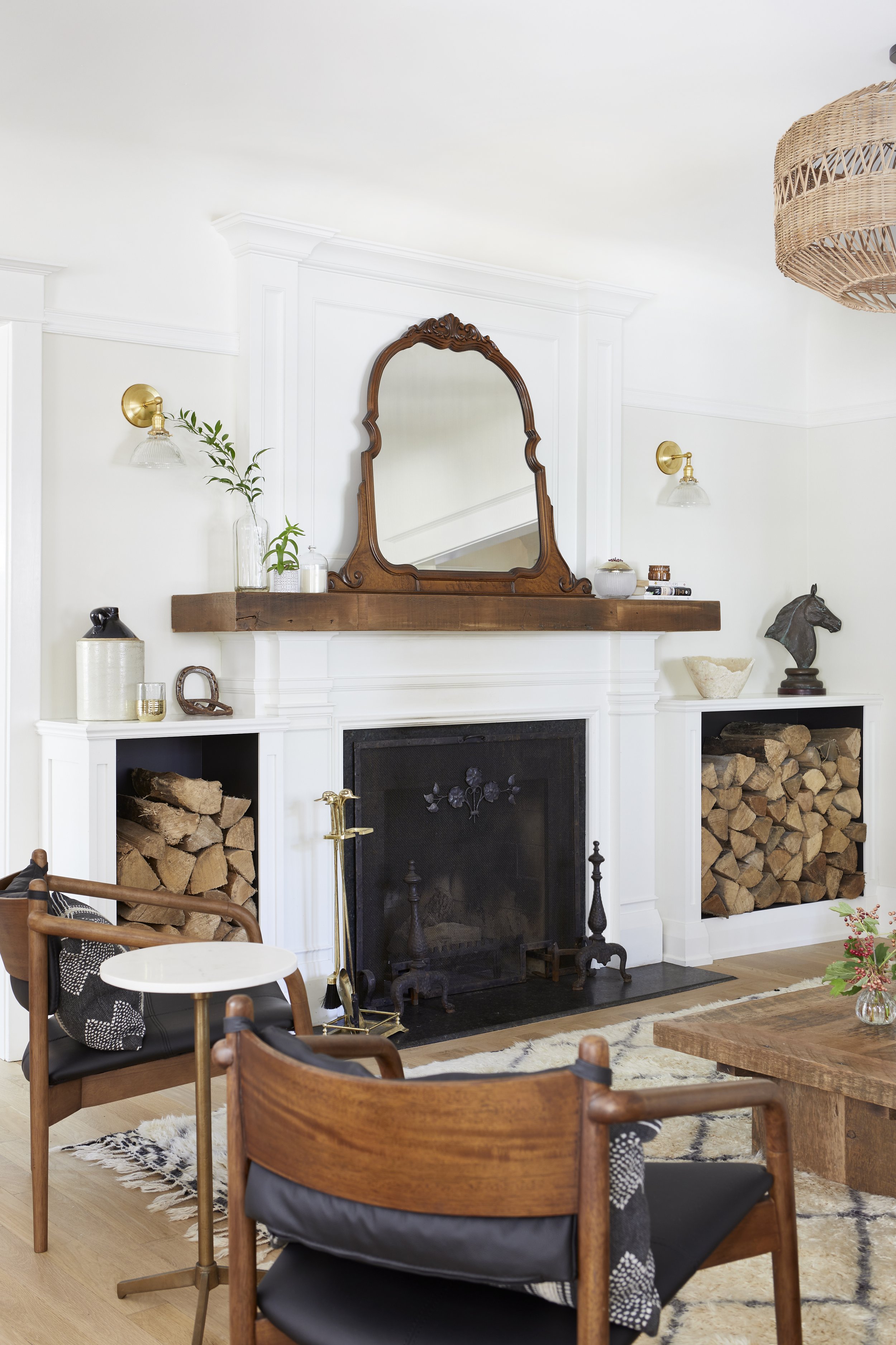 Living room with a white fireplace, wooden mantel, decorative objects, a large mirror, wood storage logs, chairs, and a wooden coffee table.