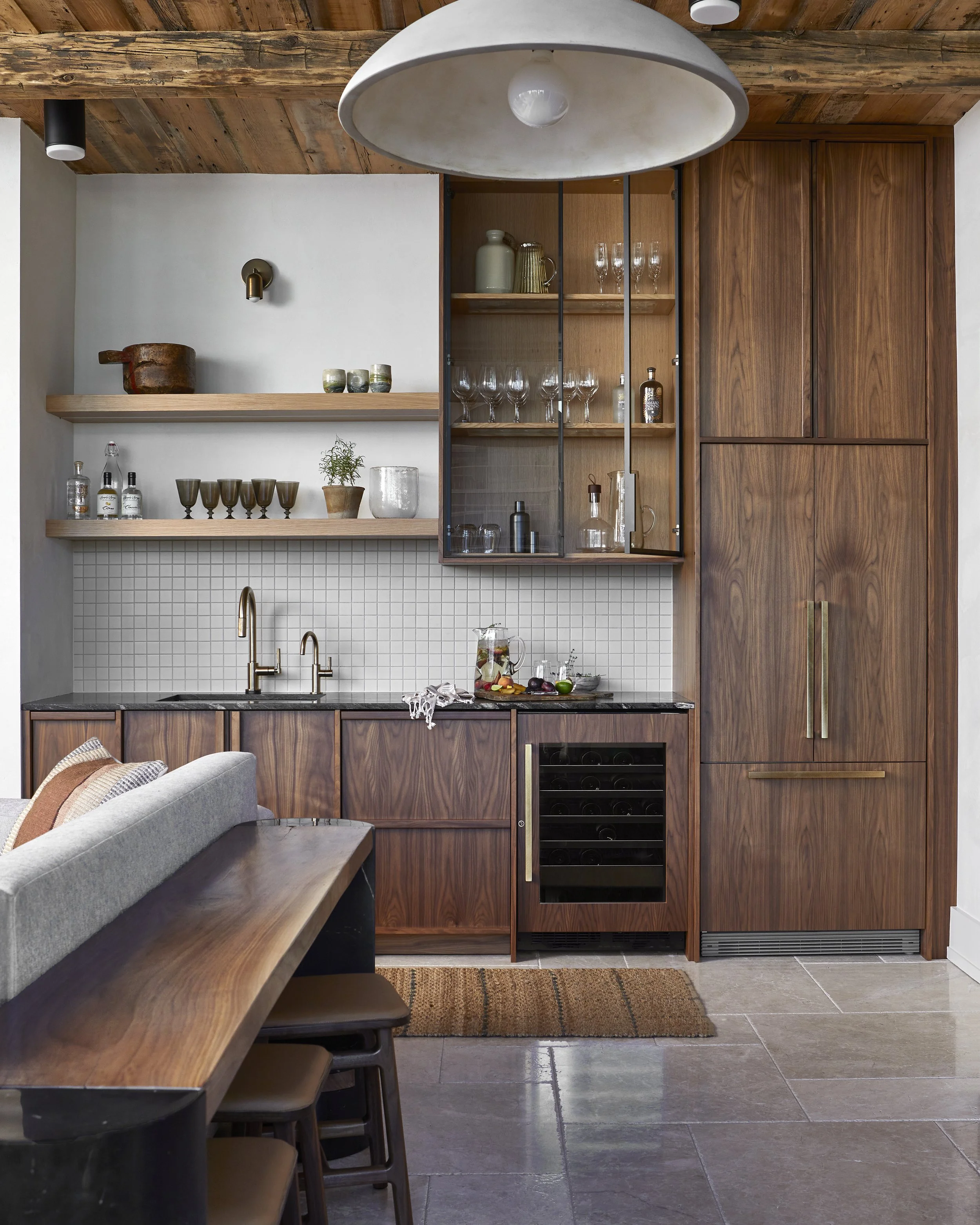 Modern kitchen with wooden cabinets, open shelves with glassware, a counter with a sink, and a built-in wine fridge. A large pendant light hangs above, and there's a small rug on the tiled floor. Part of a dining table with stools is visible in the f