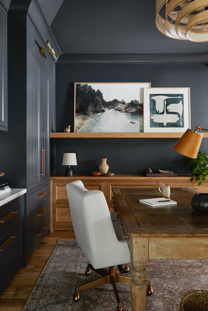 A modern home office with dark gray walls, a wooden desk, a light-colored office chair, and artwork on the wall. The room features gold accents, a large wooden chandelier, and decorative items on the shelves.