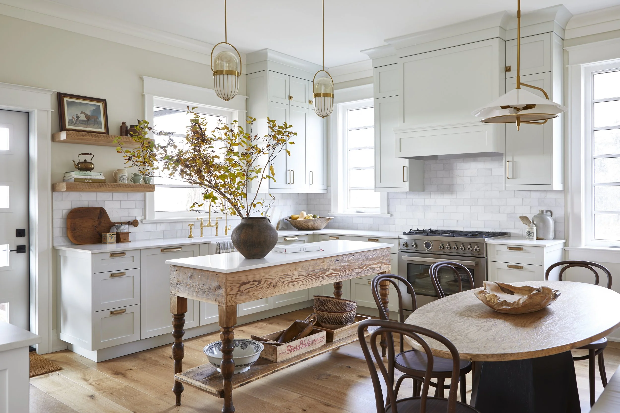 Bright and airy kitchen with white cabinets, a white countertop, and a wooden island table. Decor includes a large vase with branches, vintage bowls, and framed pictures. Large windows let in natural light, and there is a dining table with dark woode