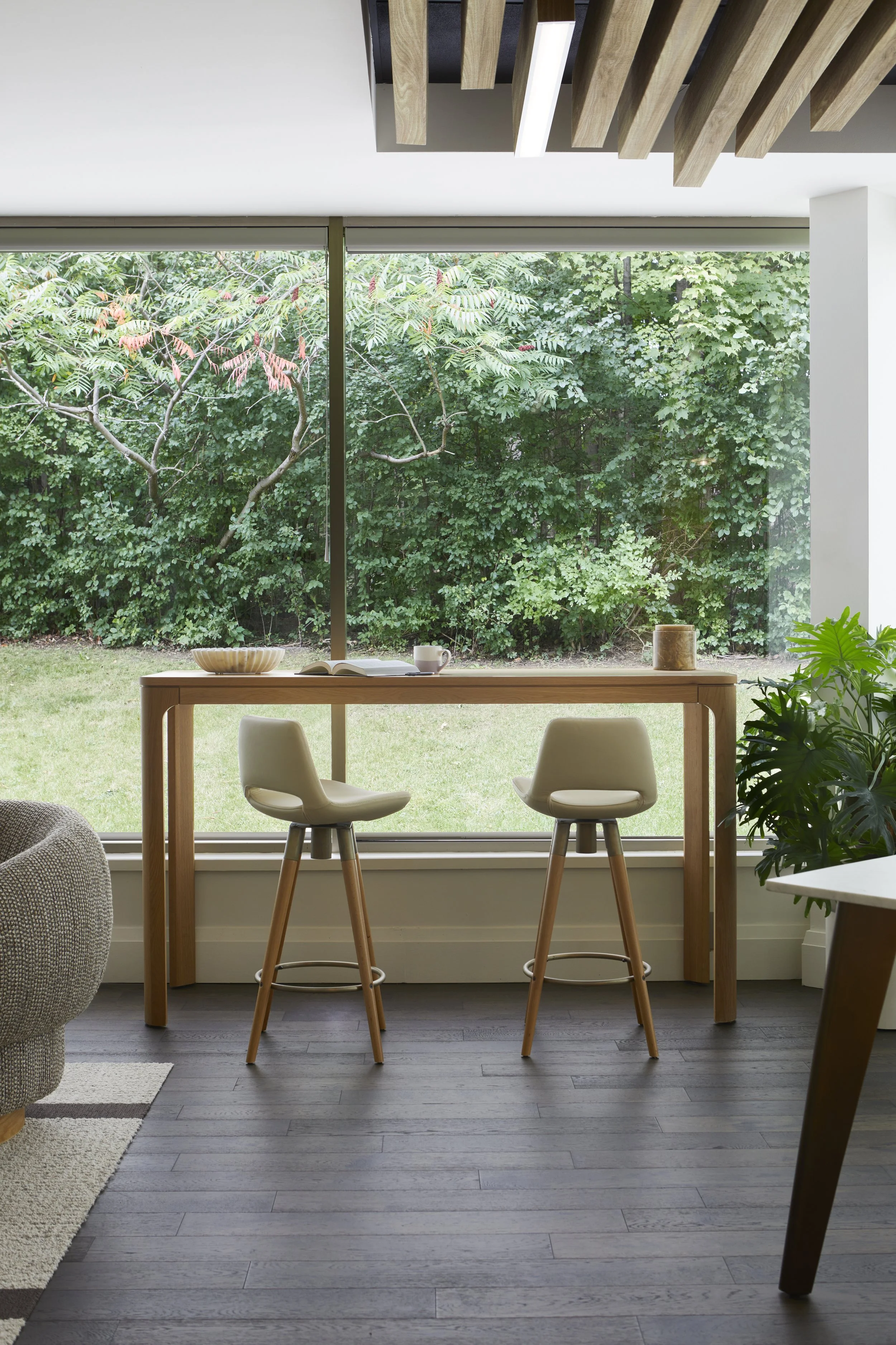Interior of a modern living room with two bar stools at a high wooden table facing a large window with a view of green trees and bushes outside. There is a gray sofa partially visible on the left and a potted plant on the right.