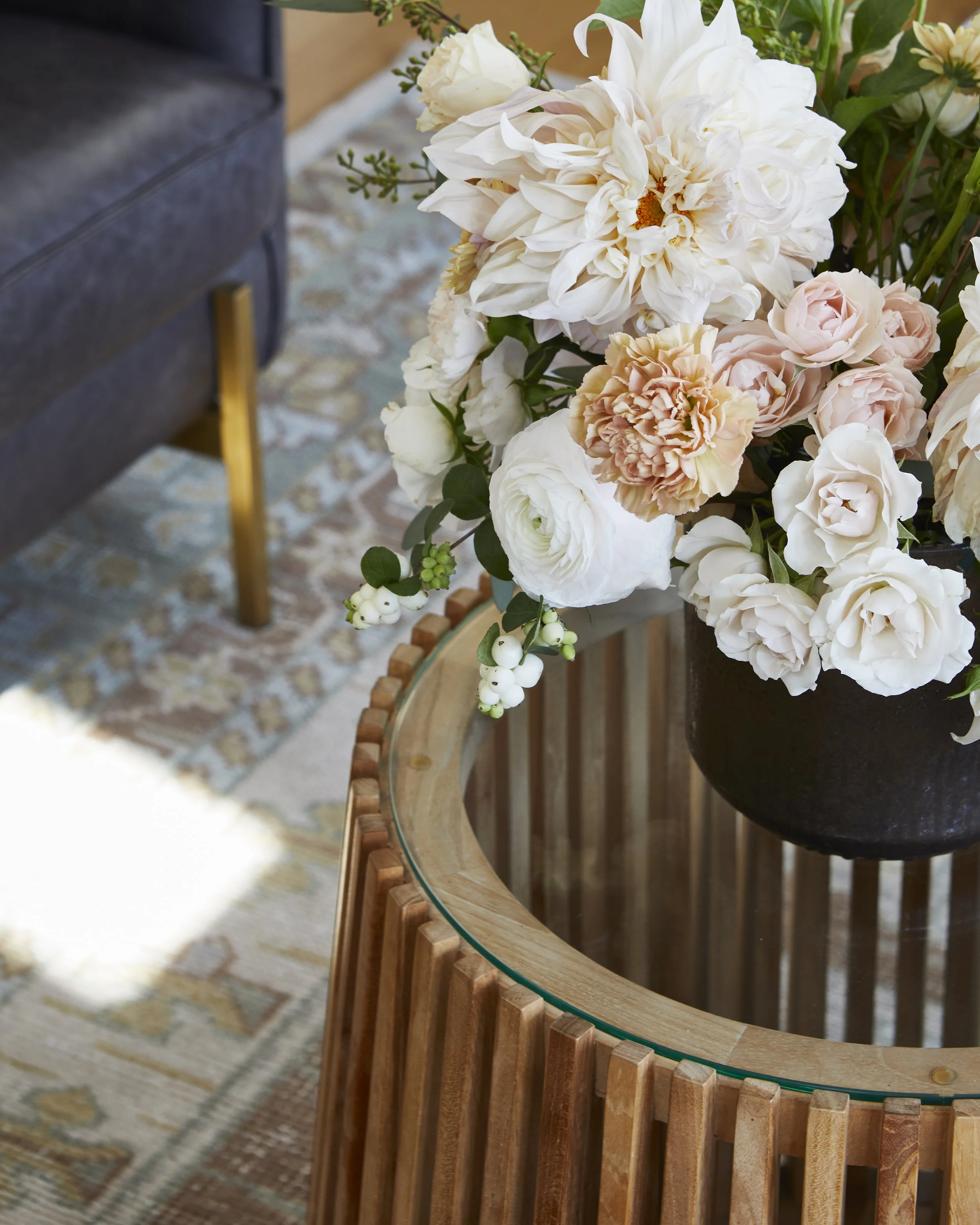 A floral arrangement with white, cream, and blush-colored flowers in a black vase on a wooden table with a glass top.