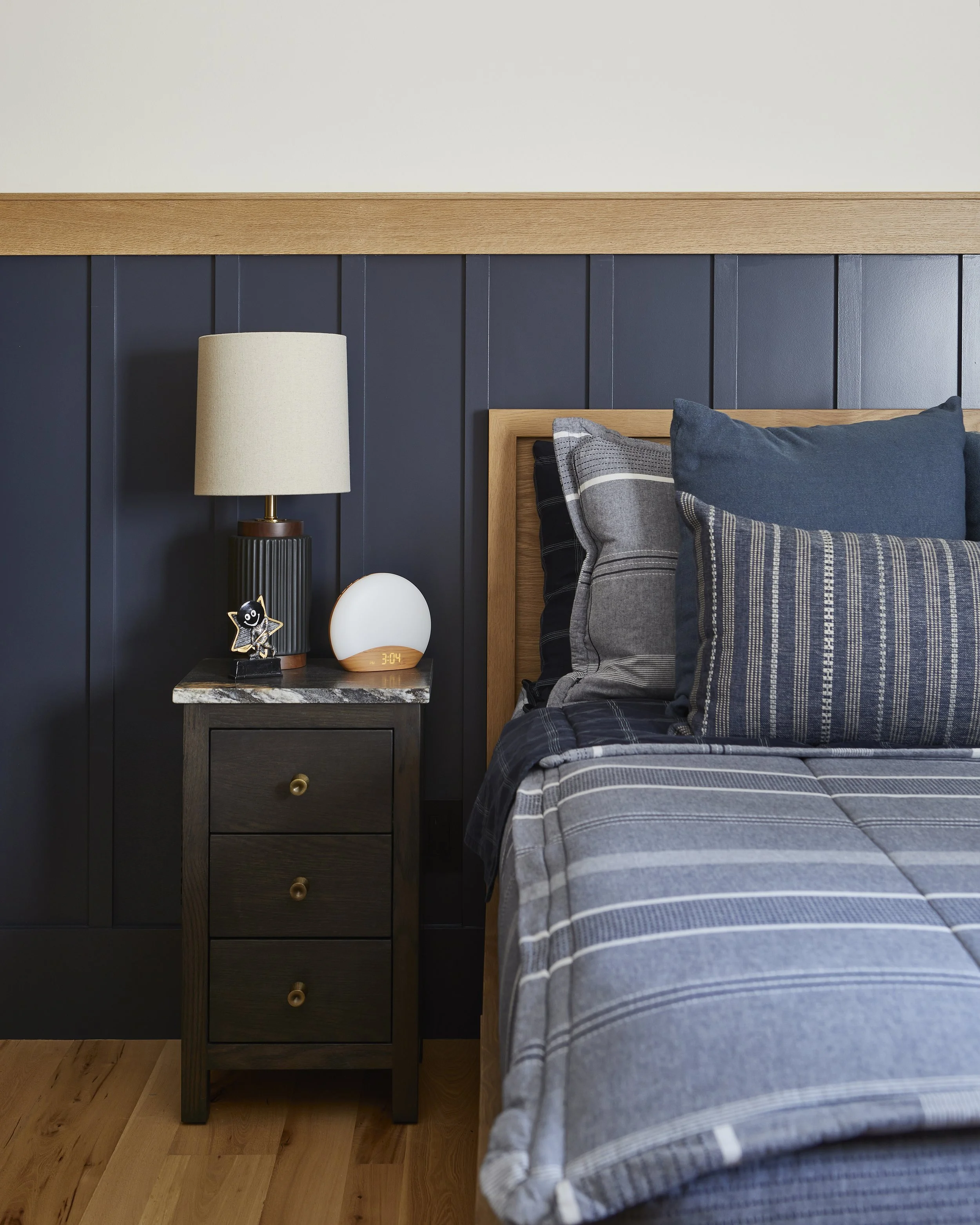 Close-up of a bedroom with a dark wooden nightstand, a lamp, a digital clock, and a decorative figurine, next to a bed with blue and gray plaid bedding and pillows, against a black wooden paneled wall.