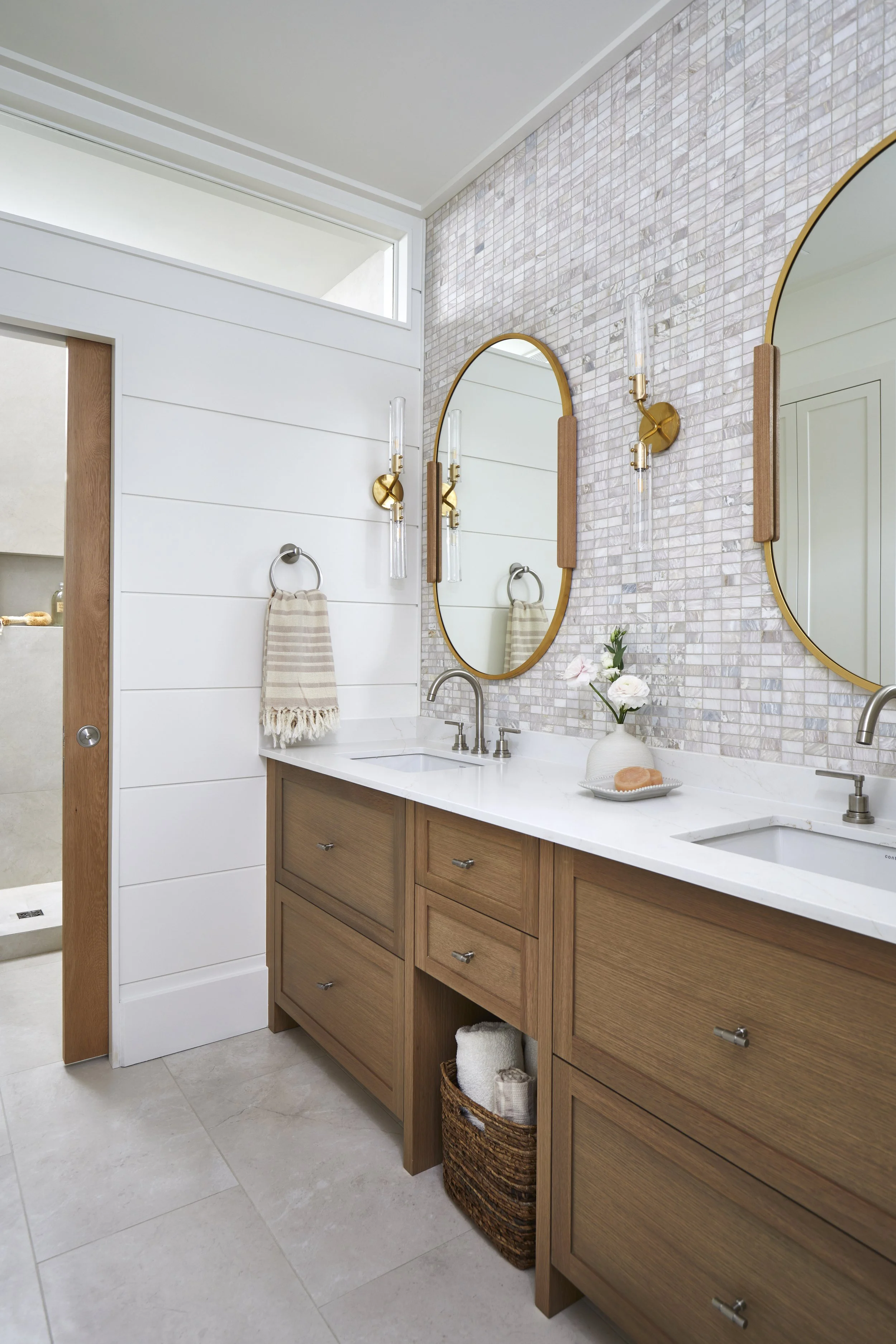 A modern bathroom with a double vanity, oval mirrors with gold frames, beige wood cabinets, and a mosaic tile wall. There are wall-mounted light fixtures with glass and gold accents, a vase with flowers, a soap dish, and towels.