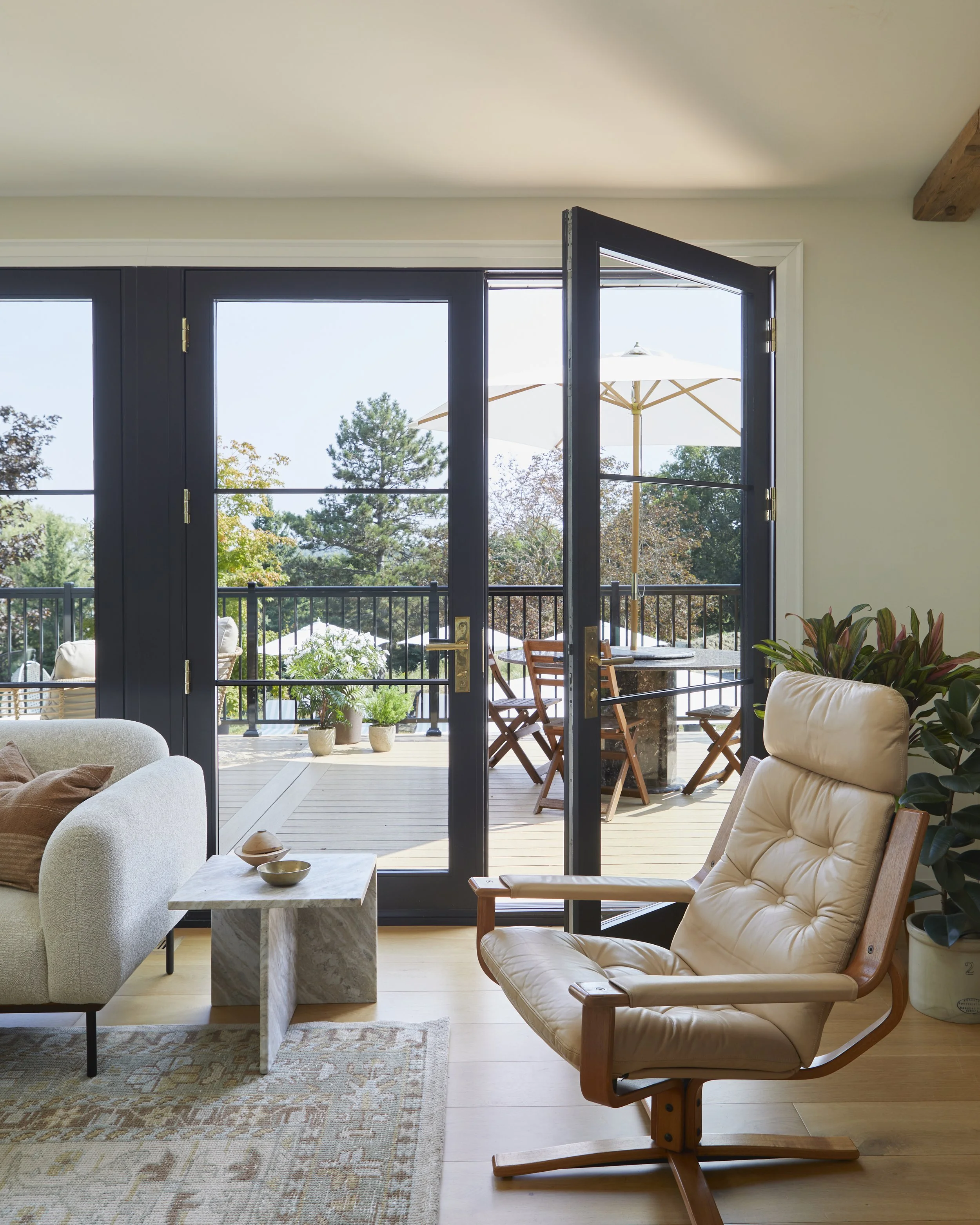 Living room with sliding glass door open to outdoor patio, showing outdoor table with chairs, umbrella, potted plants, and trees.