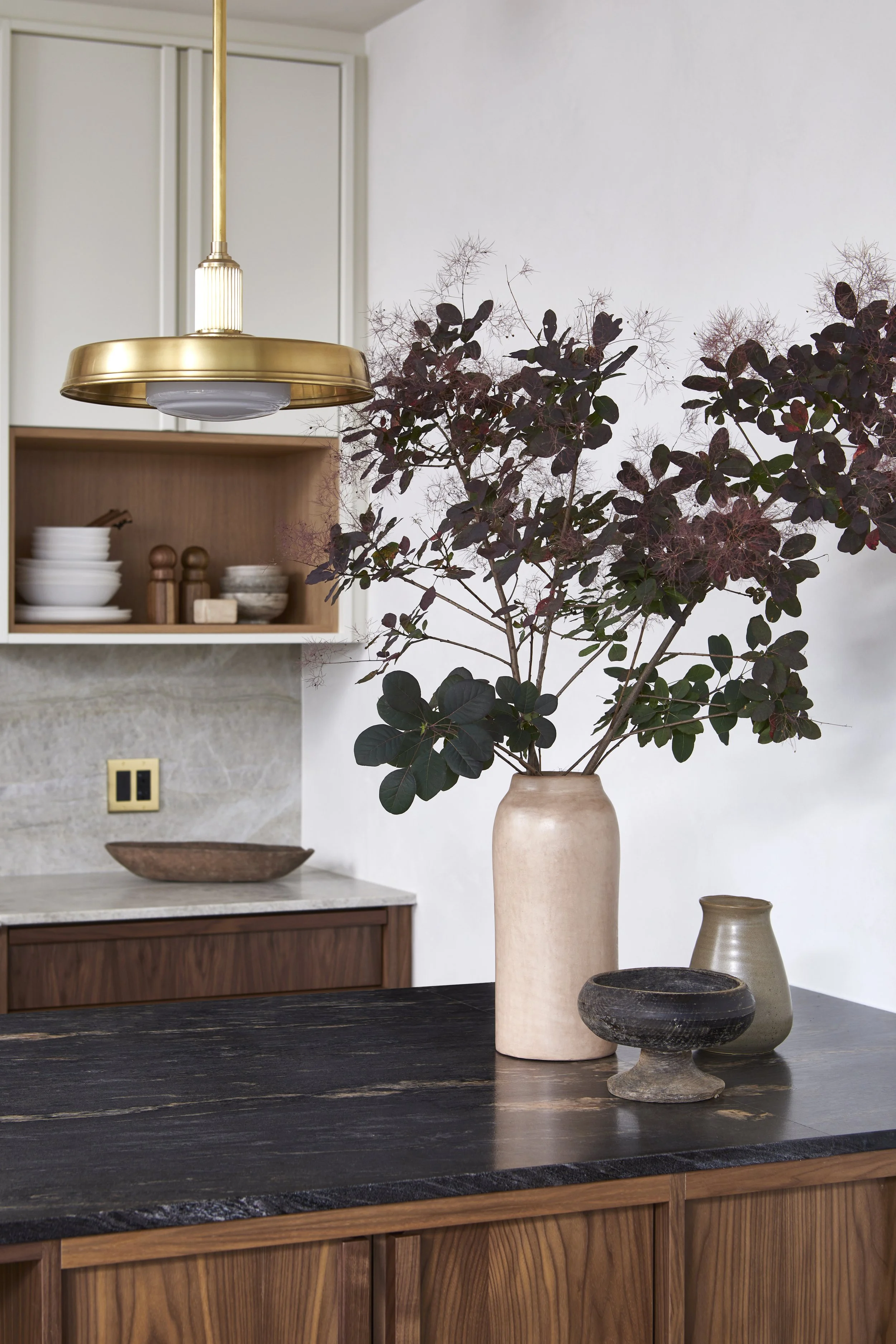 A kitchen interior with a large ceramic vase holding a bouquet of dark purple and green leaves, accompanied by two smaller vases on a dark wooden countertop. In the background, there are open shelves with bowls and jars, a marble backsplash, and a go