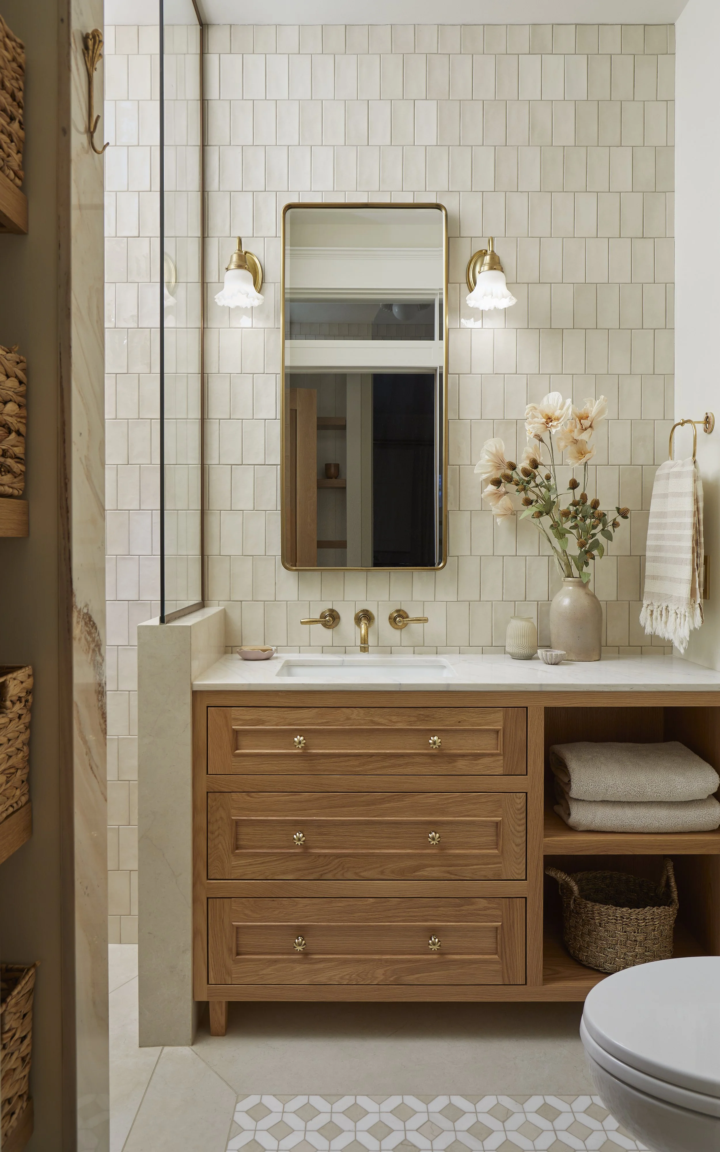 A rustic bathroom vanity with a white marble countertop, brass fixtures, a rectangular mirror, and beige tiled walls. Decor includes a vase with flowers, folded towels, and a woven basket, with part of a toilet visible in the foreground.