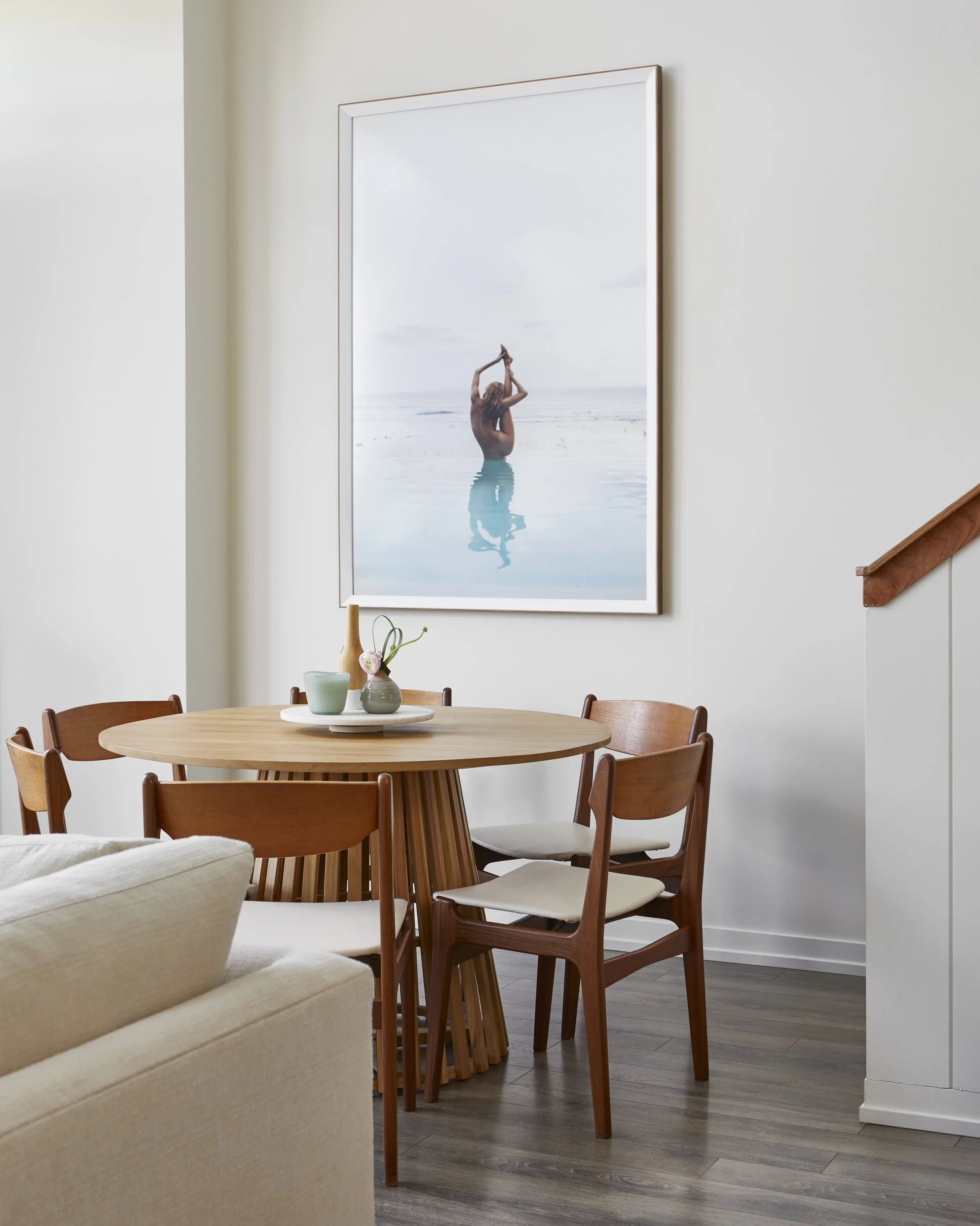 A dining room with a round wooden table and six matching chairs. On the table, there are vases and small decorative items, and a large framed picture of a woman standing in the water, performing a yoga pose, hangs on the white wall behind the table.