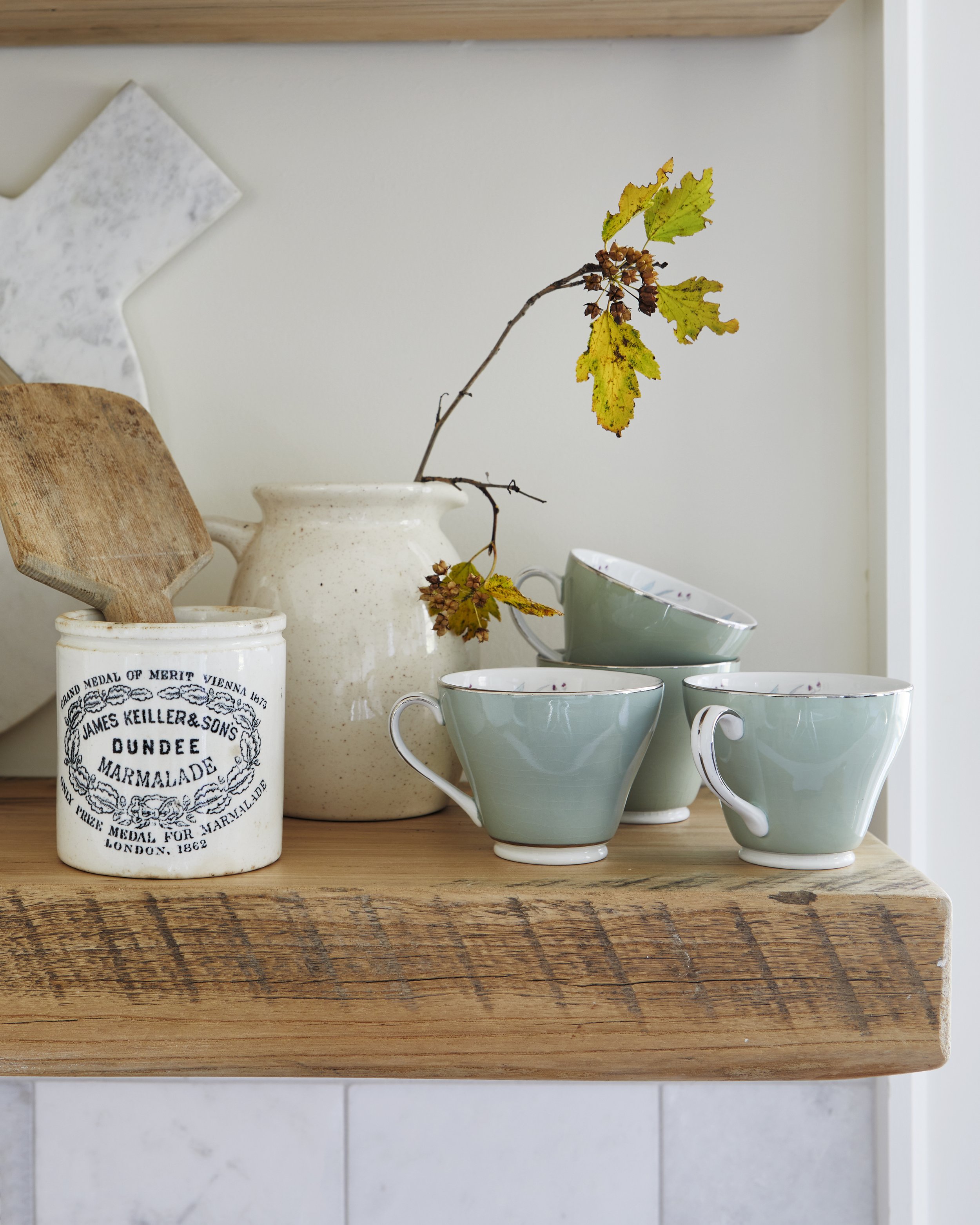 A wooden kitchen shelf with a white jar labeled 'Dundee Marmalade' holding a wooden spatula, a beige ceramic vase with a branch of yellow-green leaves, and three green teacups stacked and arranged decoratively.