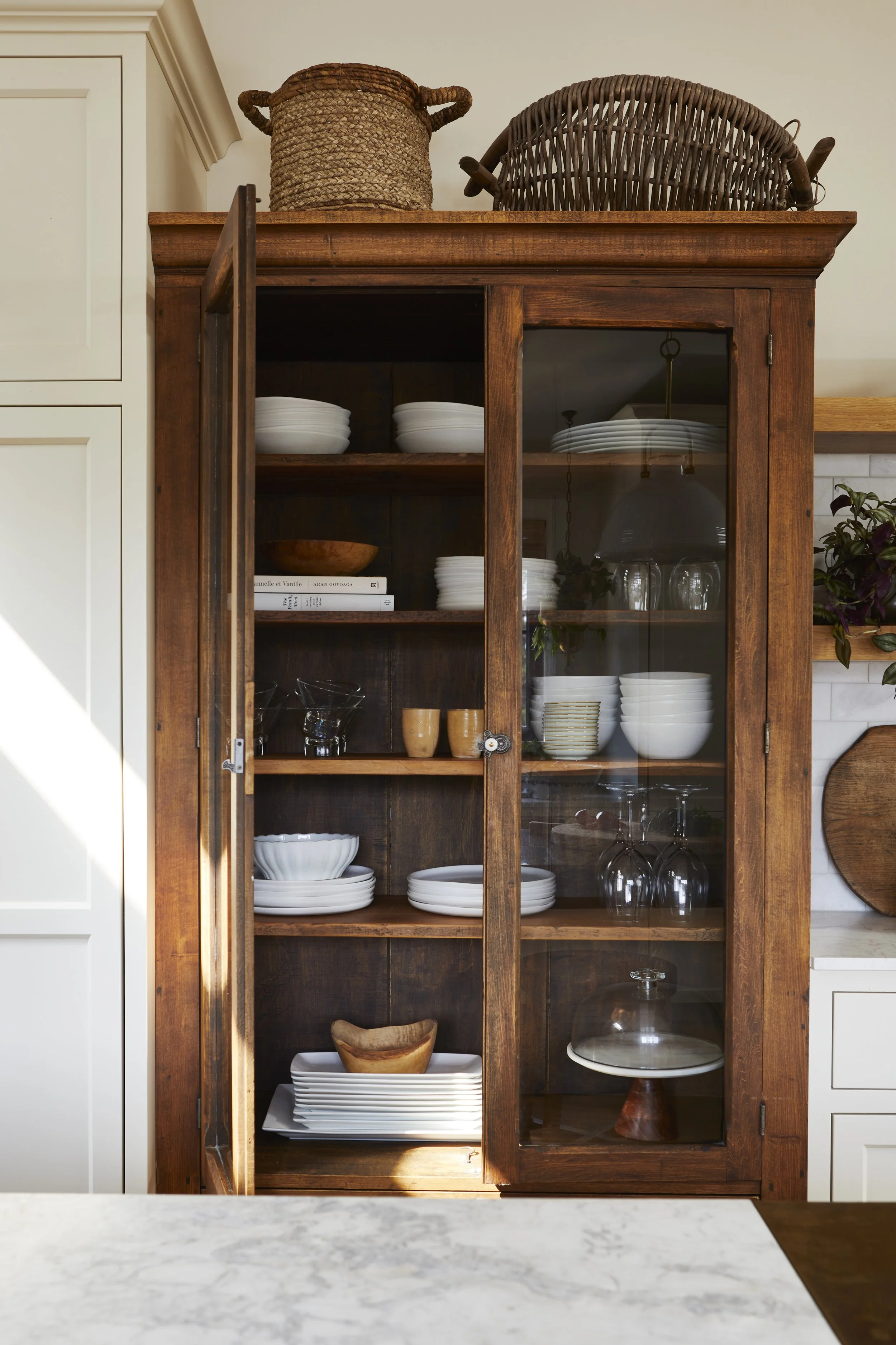 A wooden china cabinet filled with white dishes, glassware, and wooden bowls, with woven baskets on top.