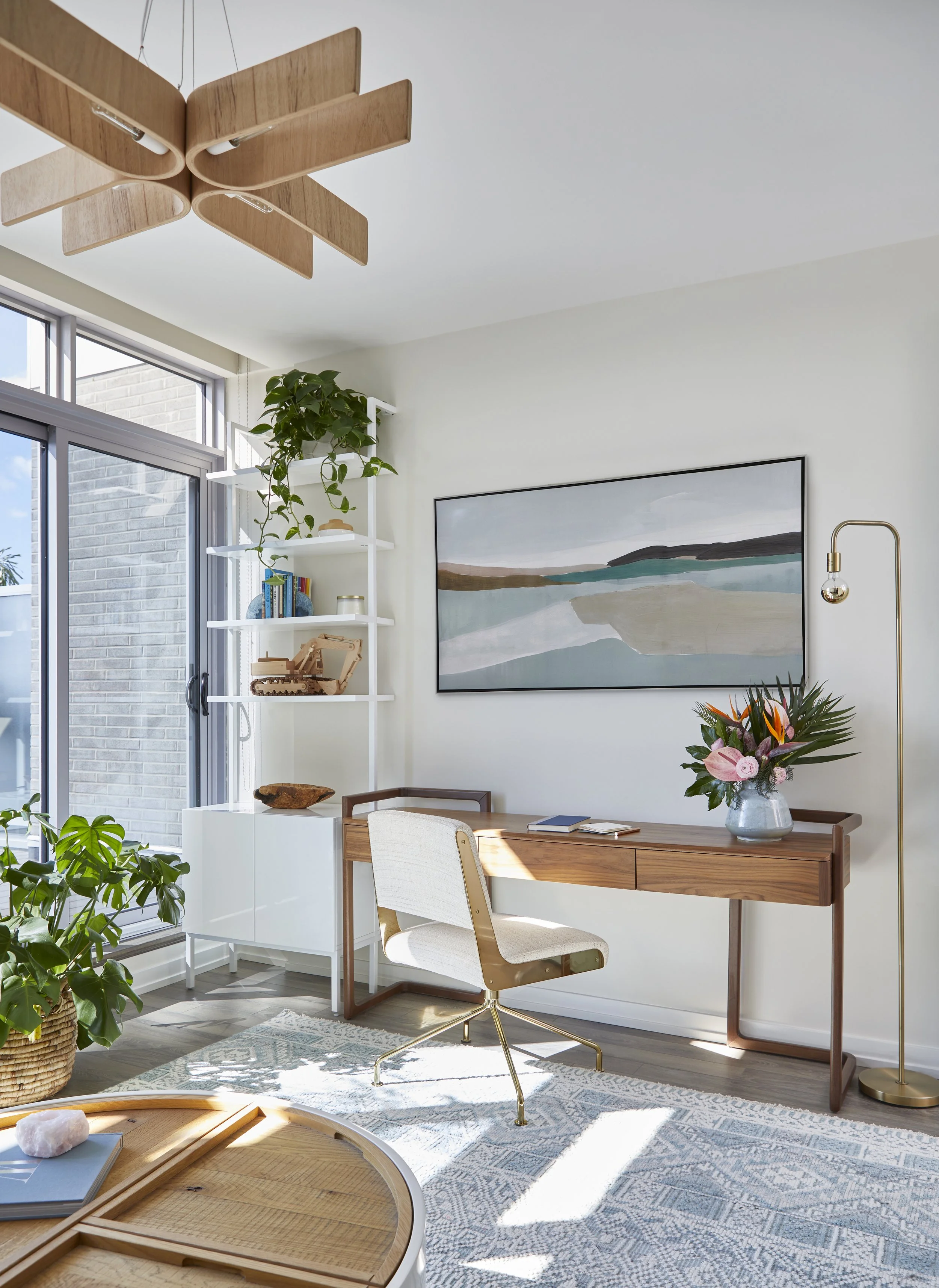 Bright and modern living or home office space with a wooden desk, white upholstered chair, a large floral arrangement, a gold floor lamp, a white bookshelf with decorative items, a large window, and abstract landscape artwork on the wall.