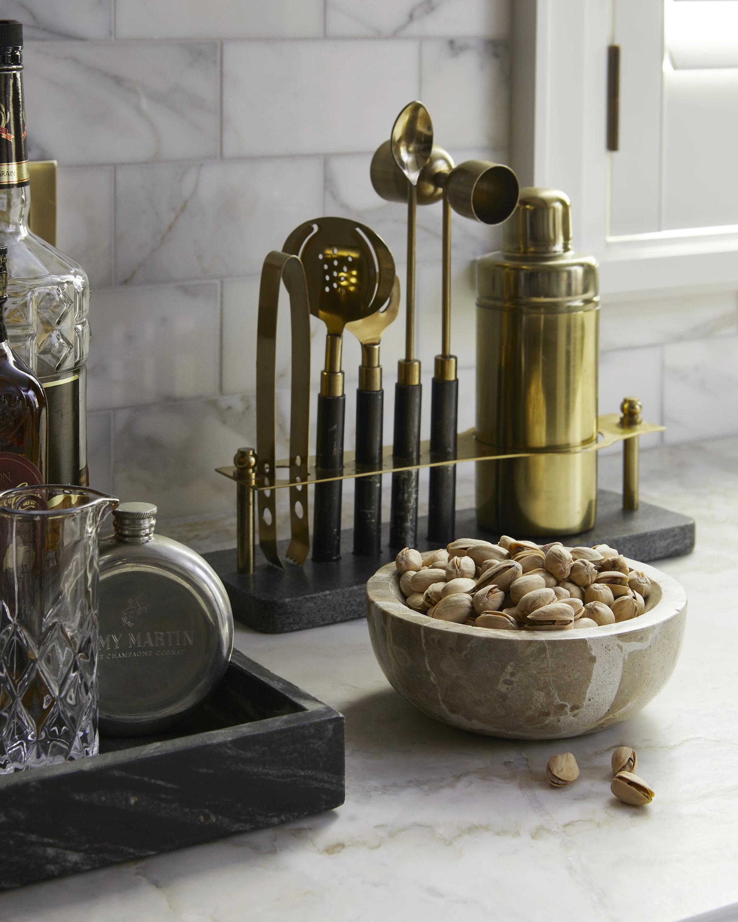 A marble kitchen countertop with a bowl of pistachios, a vintage-style flask, glass decanter, and a brass utensil holder with kitchen tools against a marble backsplash.