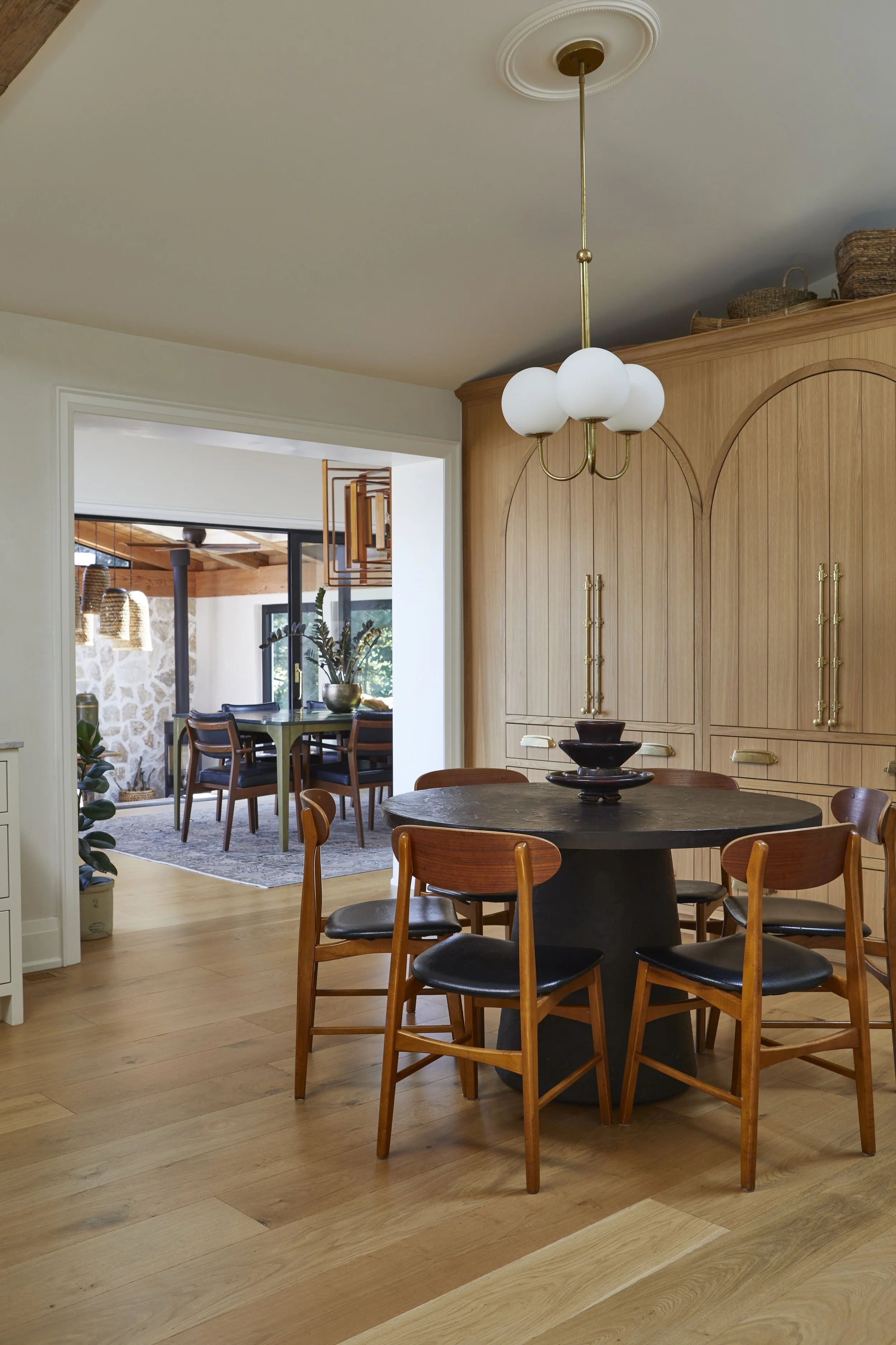 Dining room with round black table surrounded by six wooden chairs with black cushions, a modern chandelier with white globes, wood cabinetry, and visible connecting to an adjacent room with a dining area.