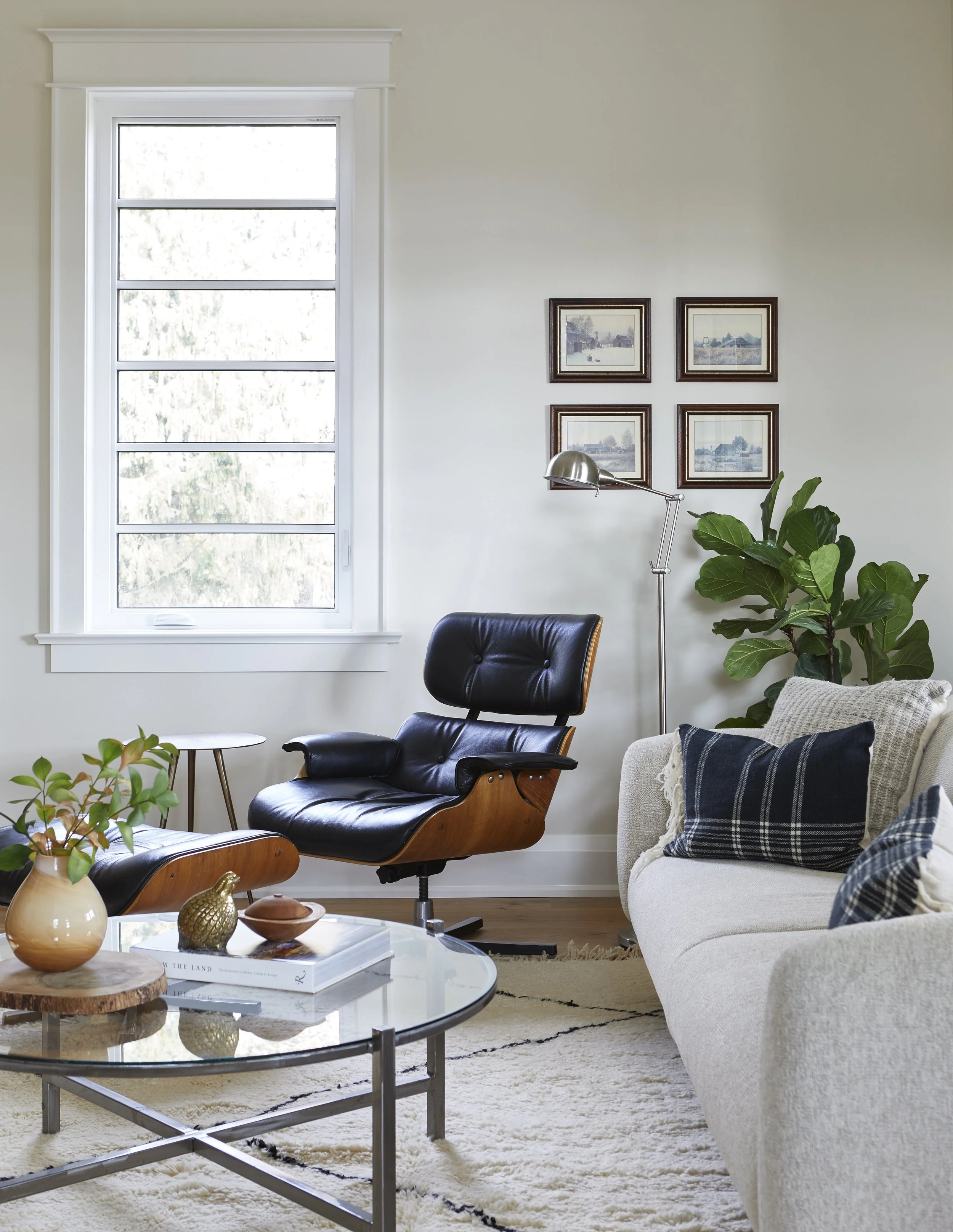 Living room with a window, black and wood lounge chair, cream sofa with plaid pillows, glass coffee table, decorative objects, a floor lamp, a large potted plant, and framed artwork on the wall.