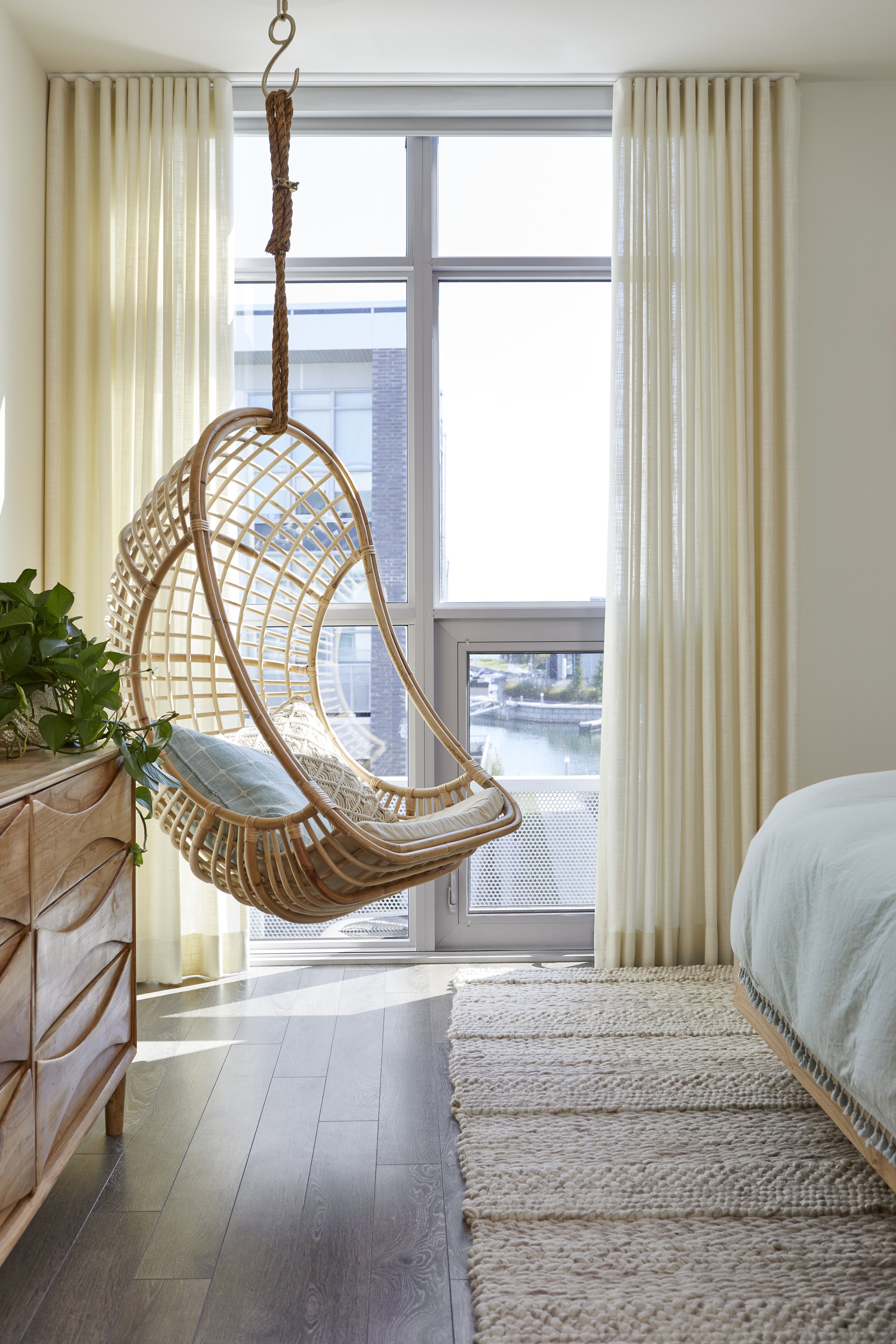 A bedroom with floor-to-ceiling windows, a wicker hanging chair, a wooden dresser with a plant, and beige curtains and rug.