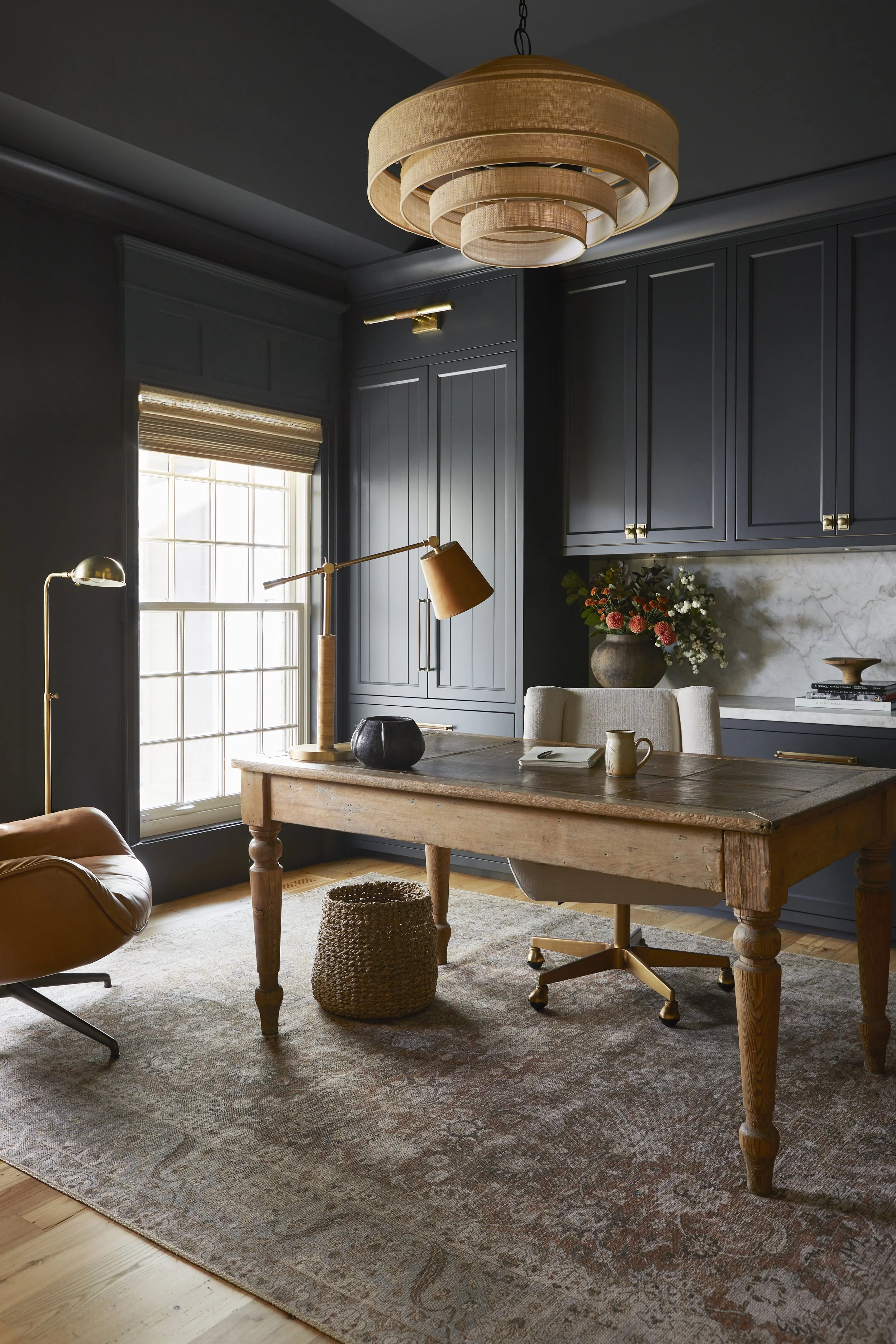 A home office with dark blue walls, a wooden desk, a beige office chair, a leather armchair, and a large window with a blind. There are decorative items like a black vase, a yellow mug, and a bouquet of flowers in a vase on the desk. A large ceiling 